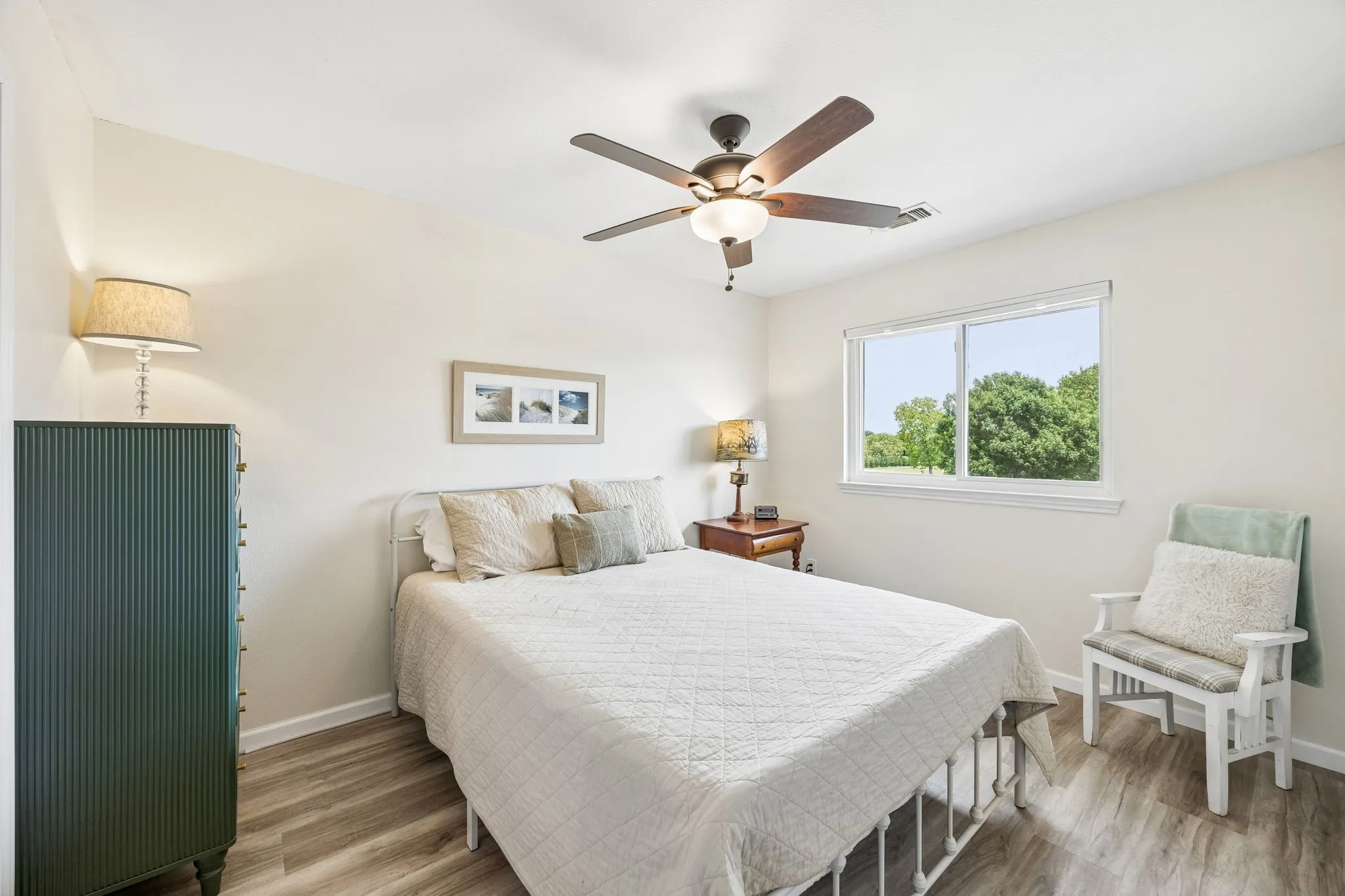 Bright room featuring wood-finish flooring, a ceiling fan with integrated lighting, and a window with exterior tree line visibility