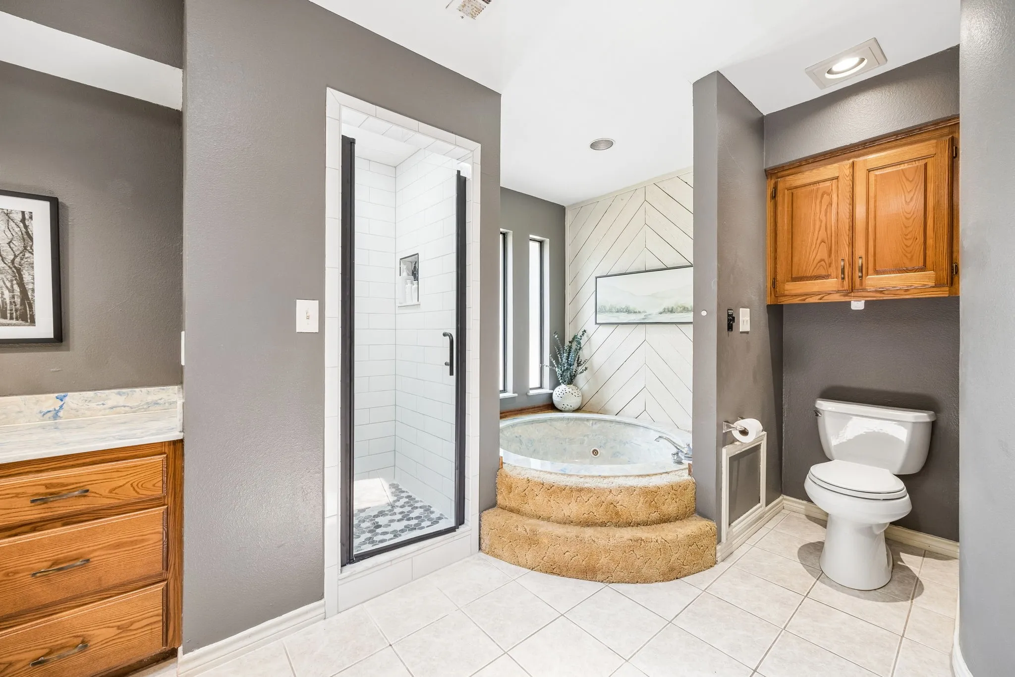 Bathroom featuring a built-in jetted tub with carpeted steps, a walk-in shower with black-framed glass enclosure, and a vanity with natural wood-finish cabinetry and a stone countertop