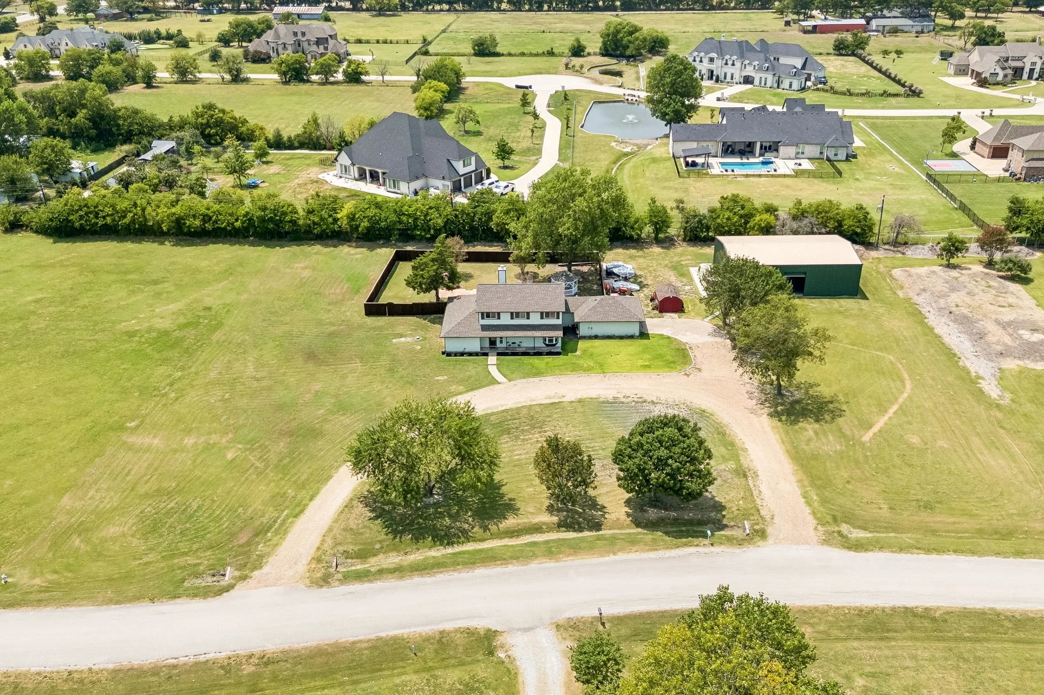 Aerial perspective showcasing a two-story residence with a light-colored exterior and dark roof, a detached green metal building, and extensive surrounding green space