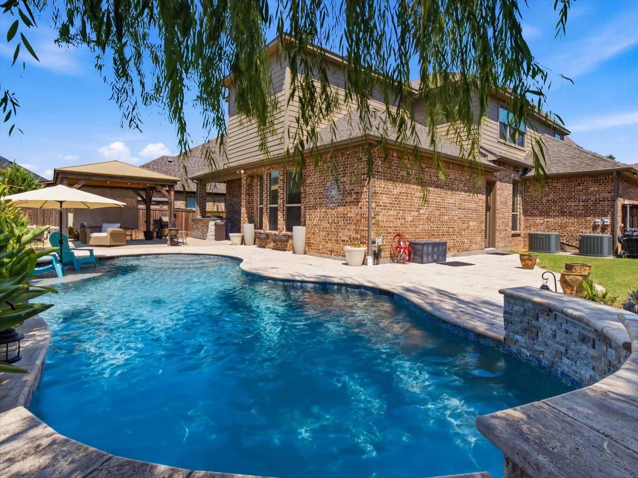 Expansive backyard featuring a freeform swimming pool, extensive paving, stone-clad retaining wall, and a separate covered gazebo structure
