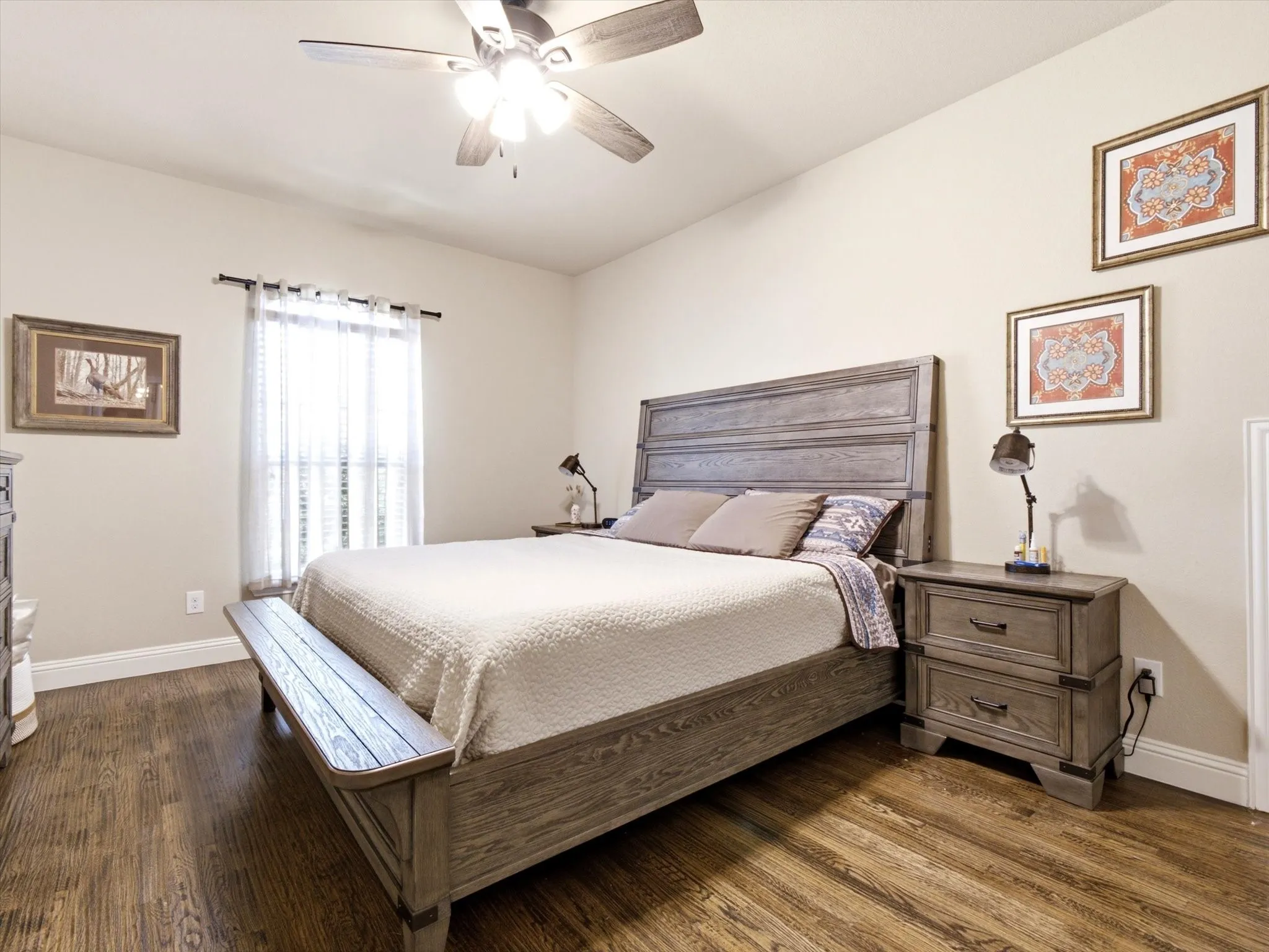 Spacious primary bedroom featuring a wood-finish platform bed with upholstered headboard, hardwood flooring throughout, ceiling fan with integrated lighting, matching wood nightstands with task lighting, and large window with sheer drapery