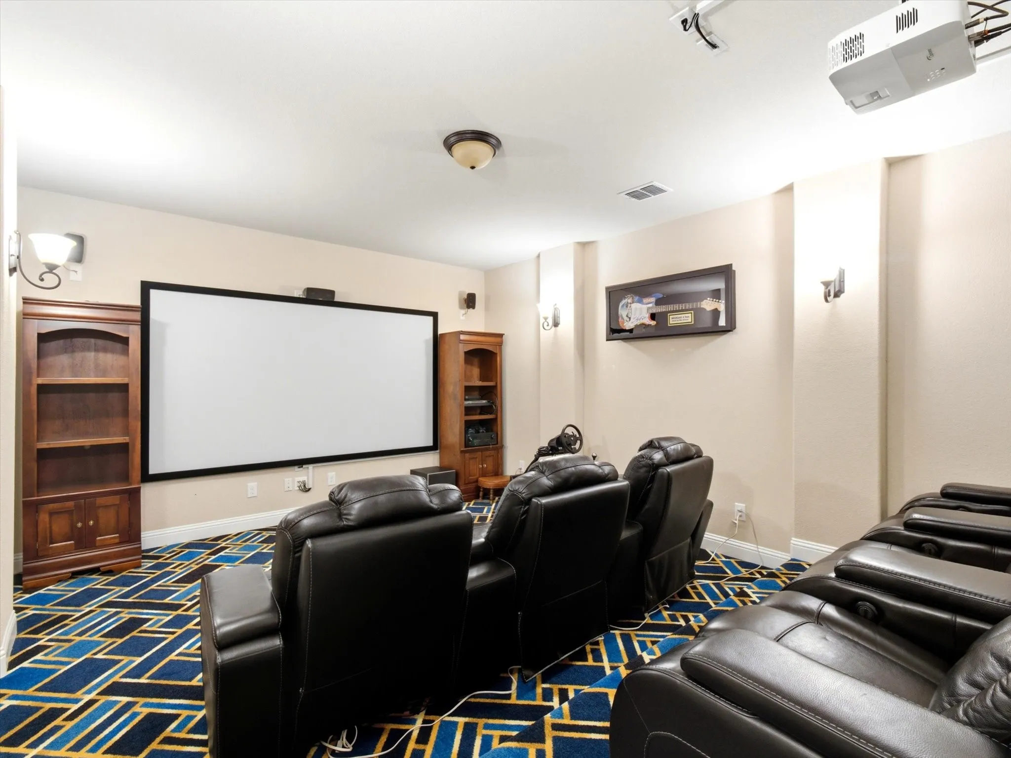 Dedicated media room featuring a large projection screen, ceiling-mounted projector, patterned carpet flooring, and built-in wood-finish shelving