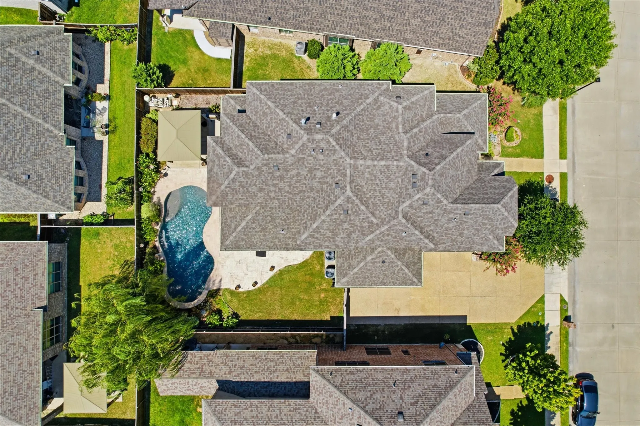 Aerial view showcasing the expansive roofline with dimensional asphalt shingle composition, in-ground swimming pool with custom blue finish, mature landscaping throughout the grounds, and substantial hardscaped patio areas