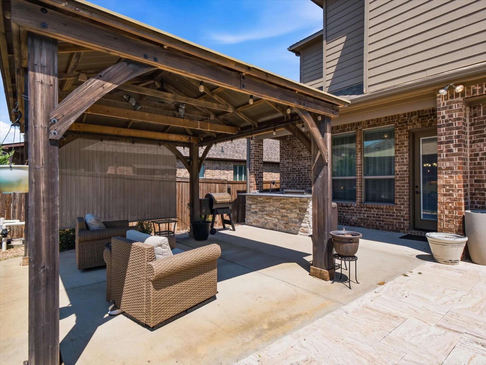 Expansive patio area featuring a wooden pergola with string lighting, a built-in outdoor kitchen with stone veneer, and a concrete patio surface