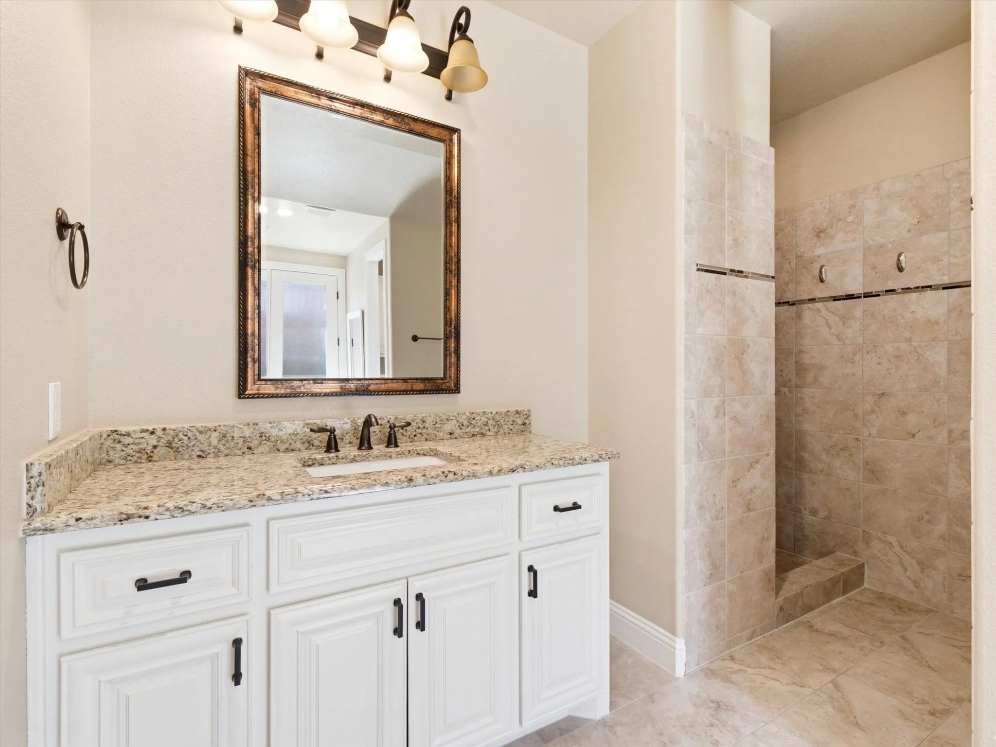 Bathroom featuring a white vanity with a light-toned granite countertop, a bronze-framed mirror, and a tiled walk-in shower with a decorative accent band