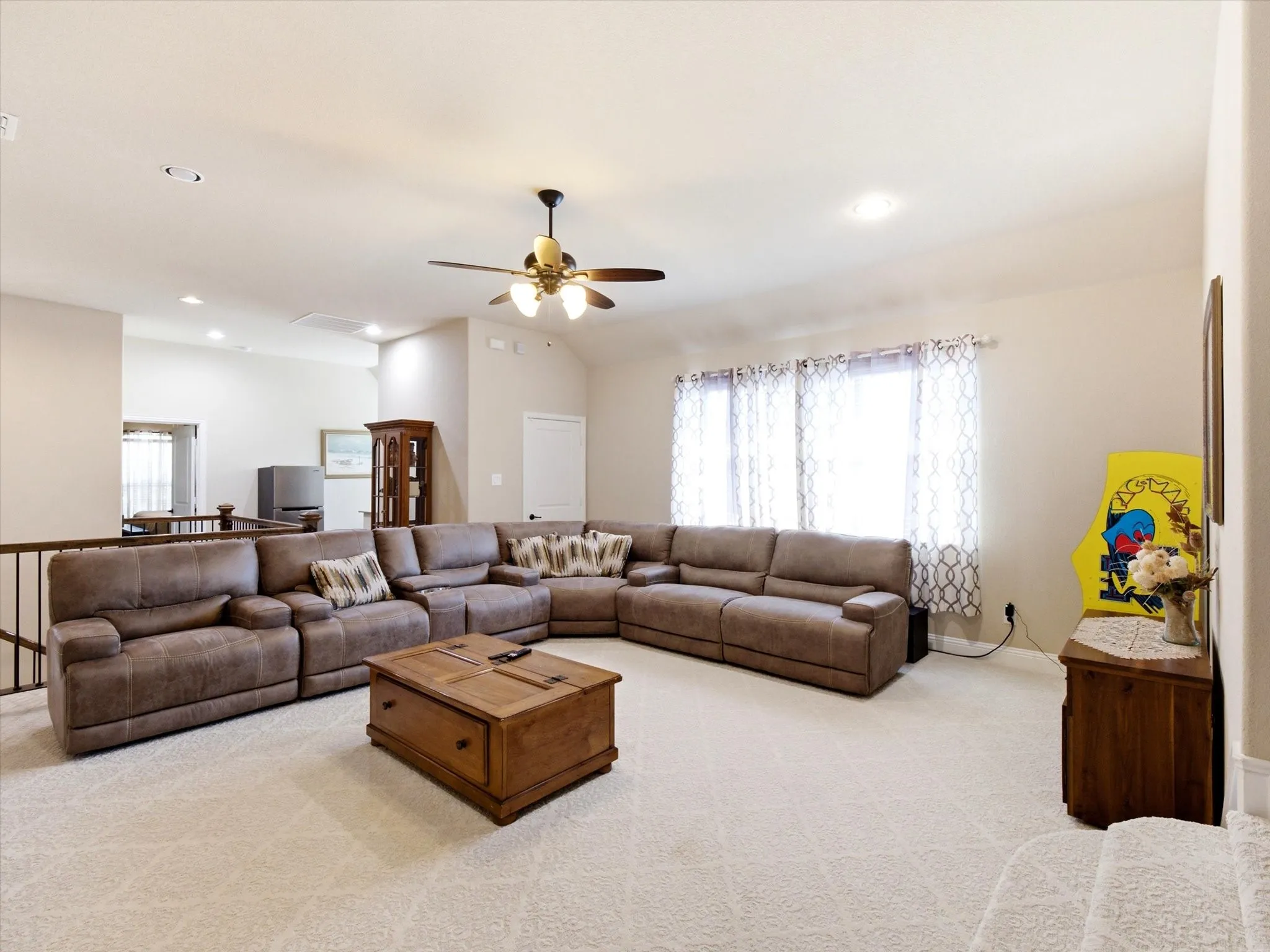 Spacious living area showcasing an expansive brown leather sectional sofa, wood-finish coffered ceiling with integrated ceiling fan and recessed lighting, neutral carpet flooring, and abundant natural light from large windows with sheer drapery