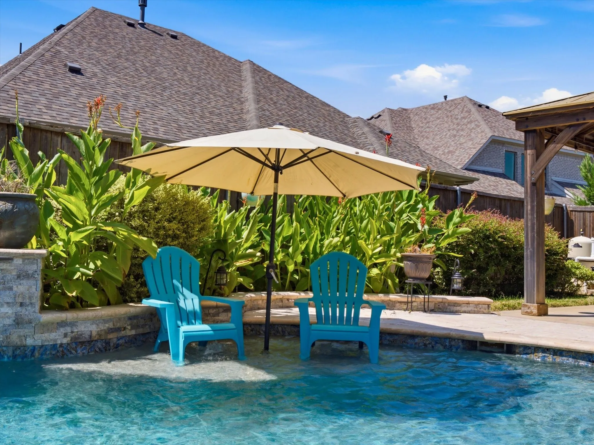 Resort-style backyard oasis featuring a sparkling saltwater pool with integrated shallow lounging areas, two Adirondack chairs positioned in the water, large cantilever umbrella providing shade coverage, mature landscaping including ornamental grasses and shrubs, and a covered pergola structure