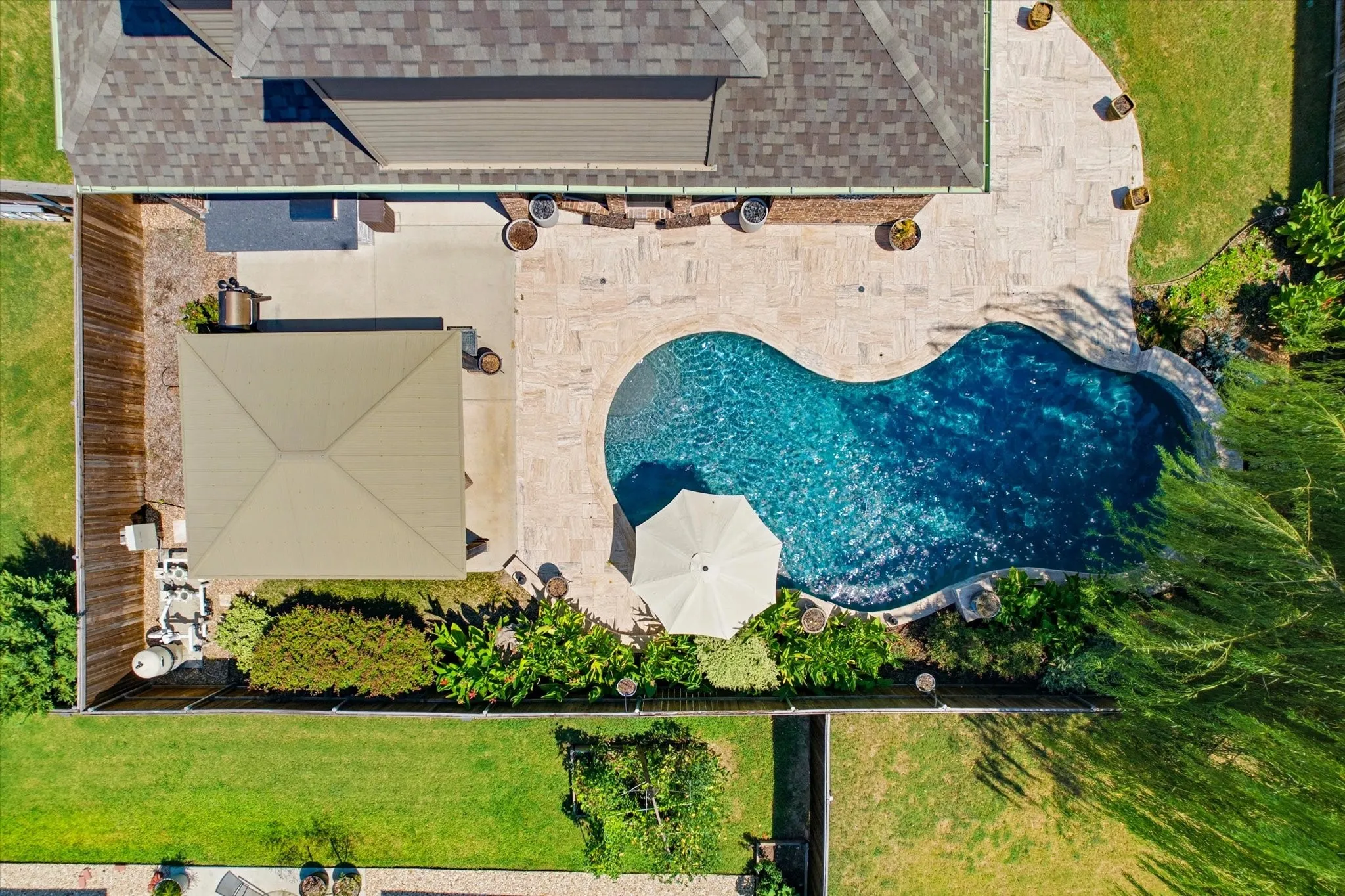 Aerial view showcasing a custom-designed free-form swimming pool with vibrant blue water, complemented by a spacious patio area featuring a retractable shade structure, manicured lawn, mature landscaping, and a brick home with architectural detailing visible in the upper portion of the property