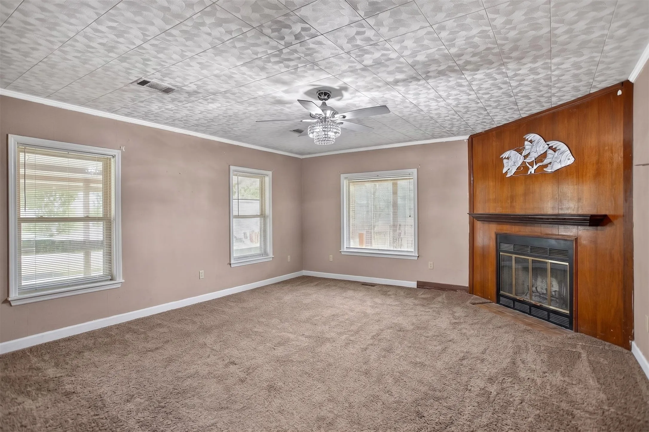 Spacious room with plush carpet flooring, multiple windows, a wood-paneled fireplace surround with a dark mantel, a decorative ceiling, and a ceiling fan with integrated lighting