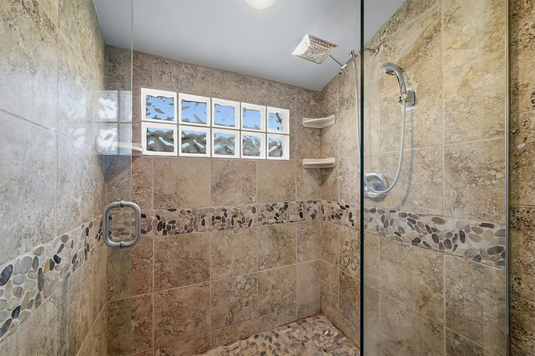 Glass-enclosed shower featuring a tiled surround, pebble tile accent band, pebble tile floor, and a glass block window