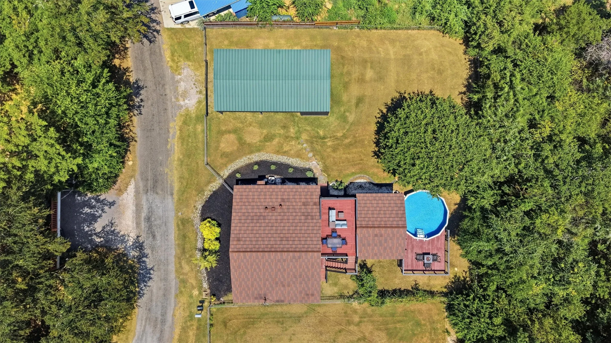 Expansive property with a brown shingle roof, a circular pool with a surrounding deck, a detached green metal roof building, and mature surrounding trees