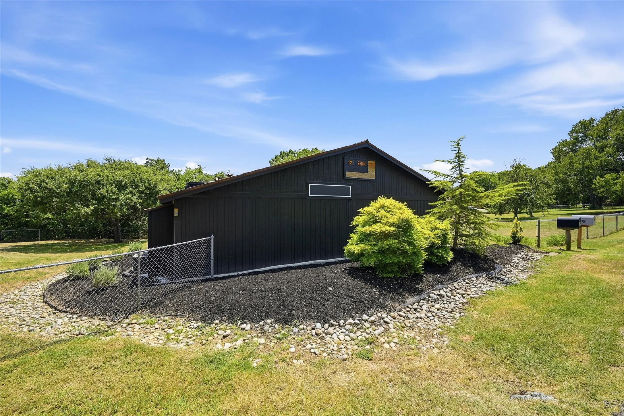 Black-paneled building with a low-slope roof, featuring a landscaped bed with dark mulch and decorative rocks