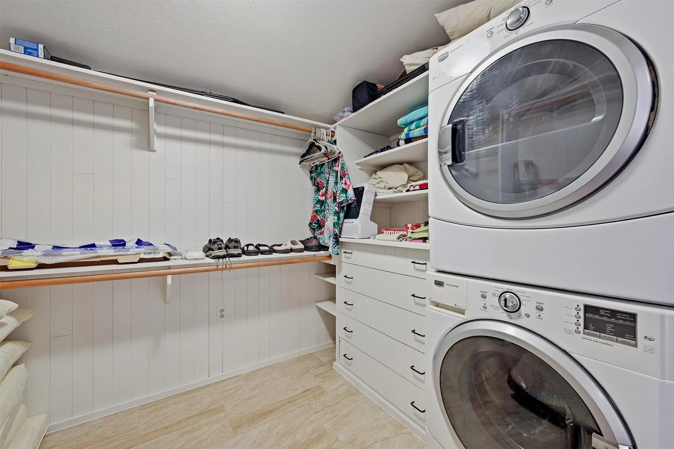 Dedicated laundry room featuring stacked front-load washer and dryer with white finish, wood-finish countertop with storage, white subway tile wainscoting, abundant cabinetry and shelving, and light wood flooring throughout