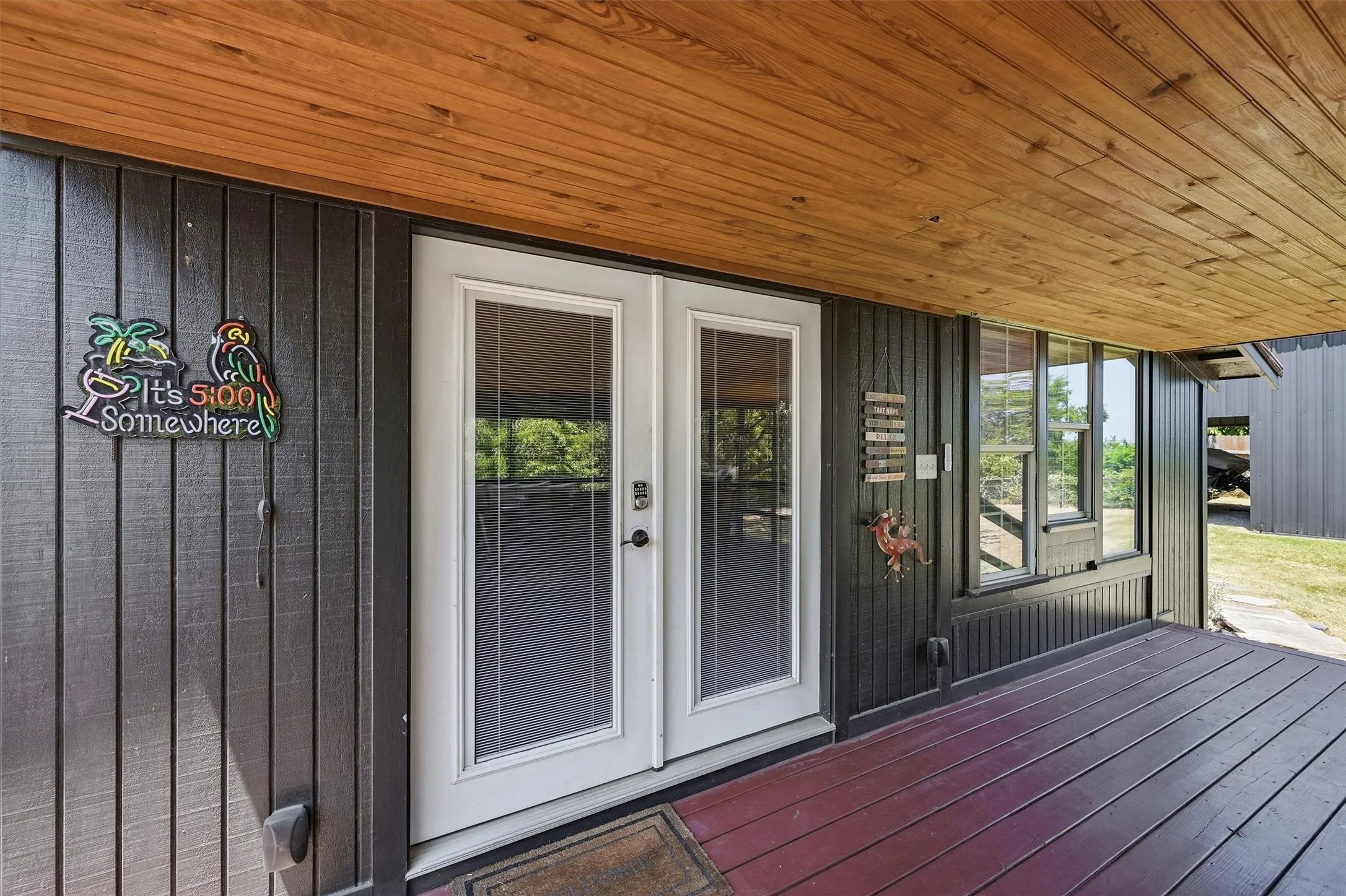 Covered porch with wood plank ceiling, double entry doors with integrated screens, dark vertical panel siding, and a red-toned wood plank deck