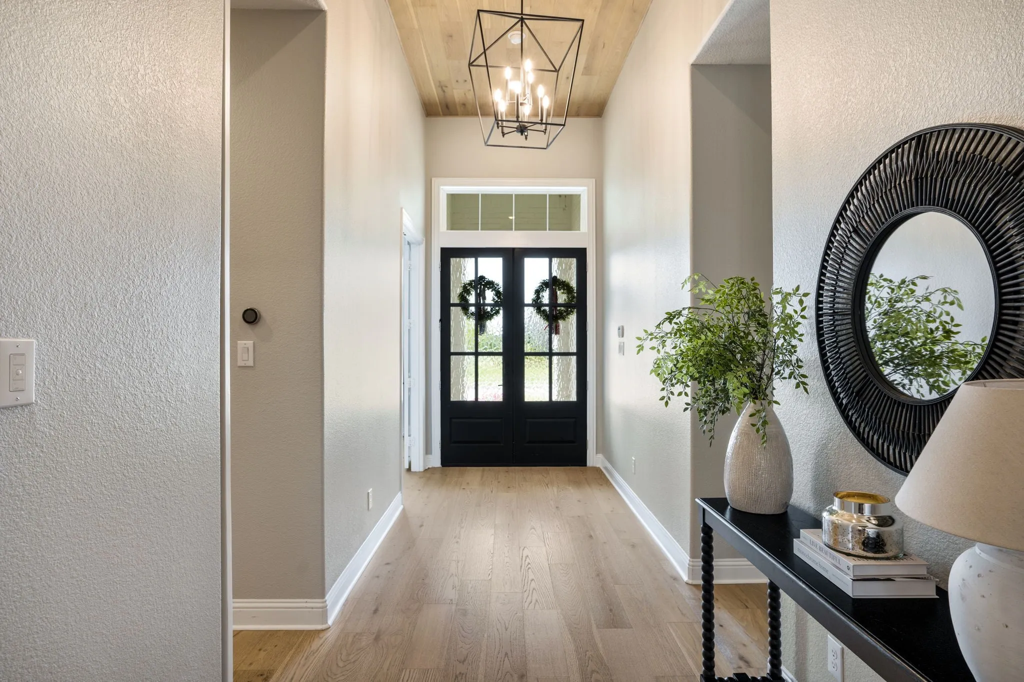Welcoming entry foyer featuring elegant black double doors with decorative glass panes, wood-finish flooring, geometric pendant lighting, and neutral wall finishes accented by taupe-toned architectural columns