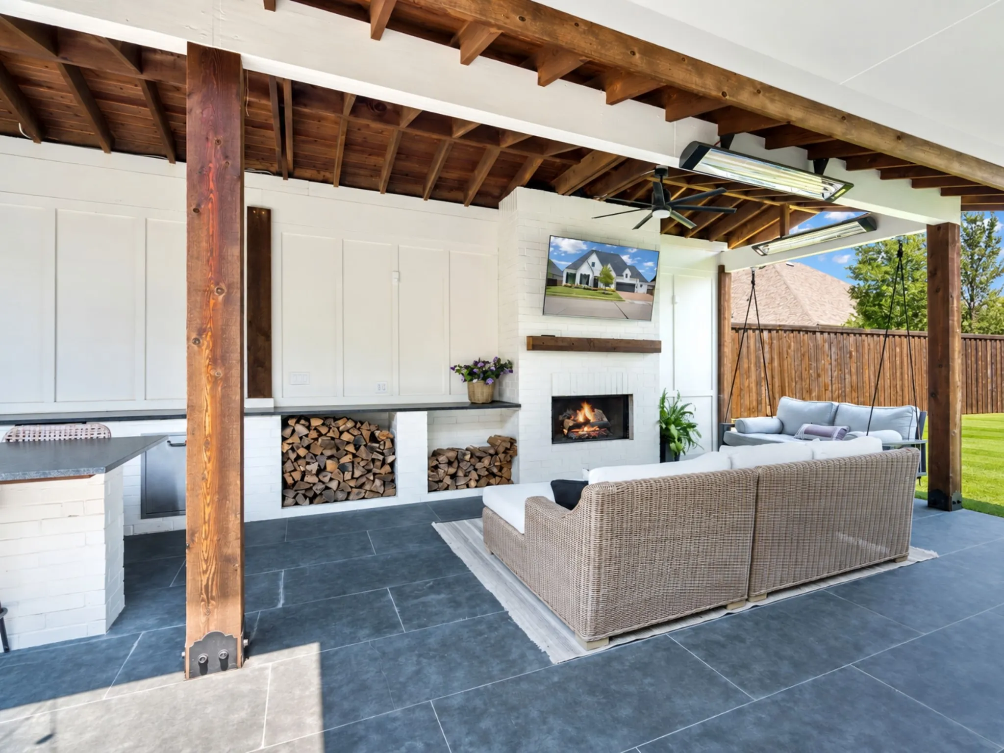 Covered patio featuring a white brick fireplace, exposed wood-beam ceiling, slate tile flooring, built-in firewood storage, and overhead heating elements