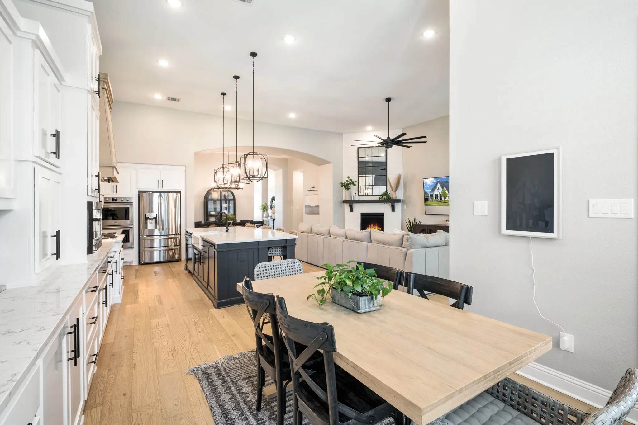 Open-concept kitchen and dining area featuring white cabinetry with stainless steel appliances, a large center island with seating, wood-finish flooring throughout, pendant lighting, and a spacious dining table