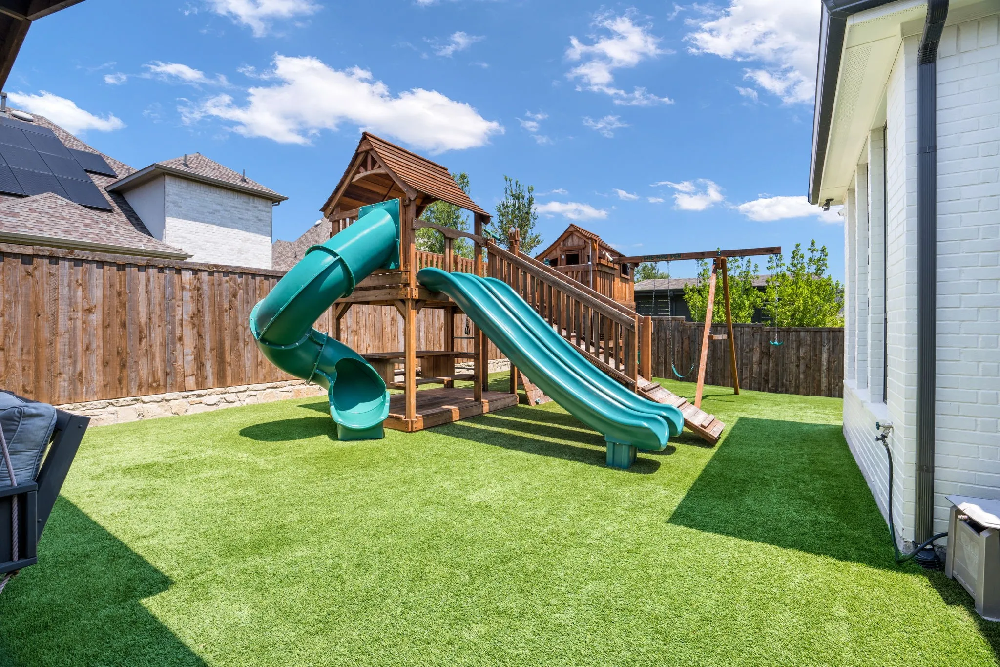 Expansive backyard oasis featuring lush, manicured lawn with a wooden-frame play structure equipped with turquoise spiral and straight slides, climbing elements, and multiple levels of decking