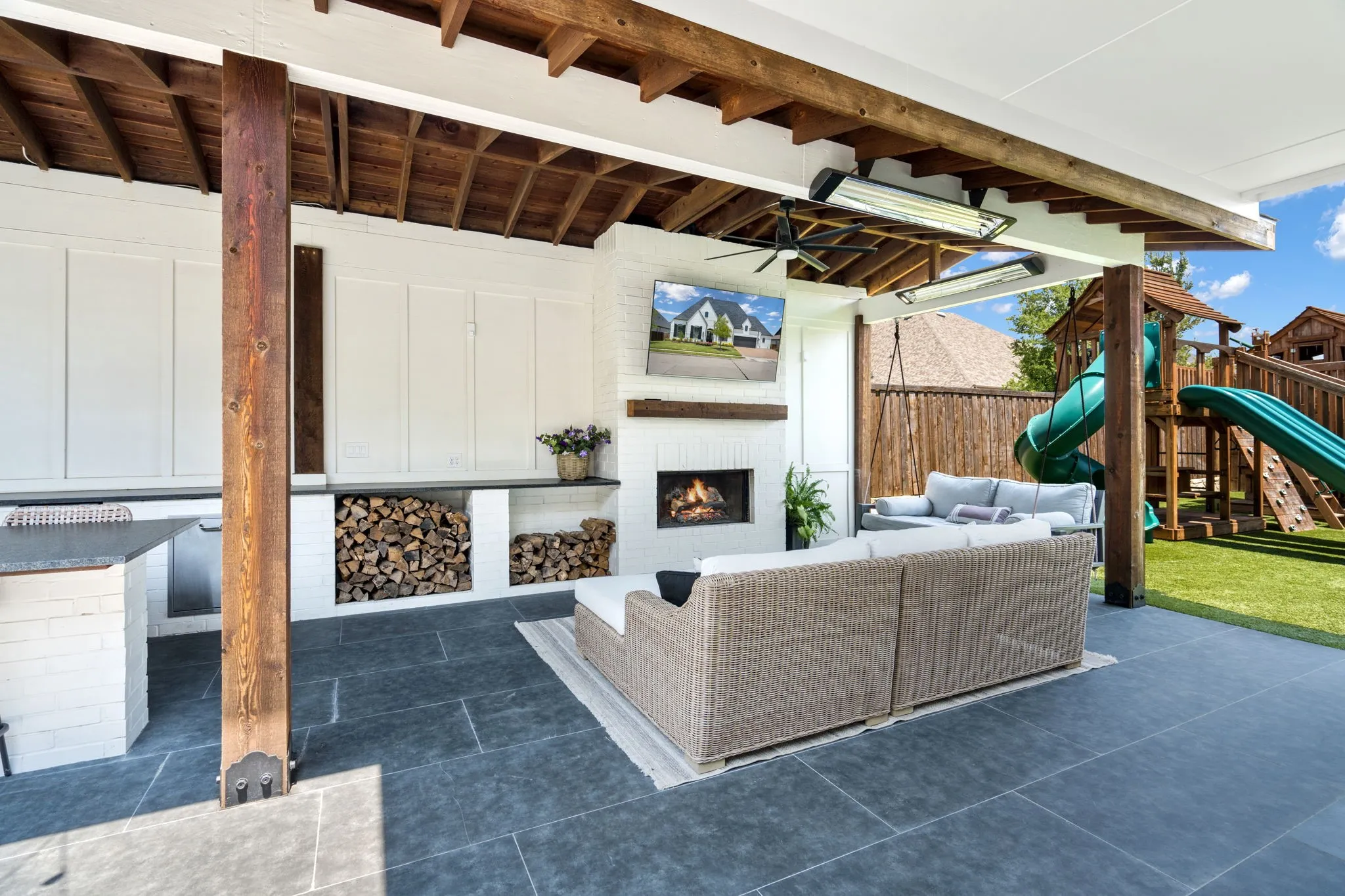 Covered outdoor living area featuring a stone tile floor, built-in fireplace with a wood mantel, paneled cabinetry, and integrated wood storage