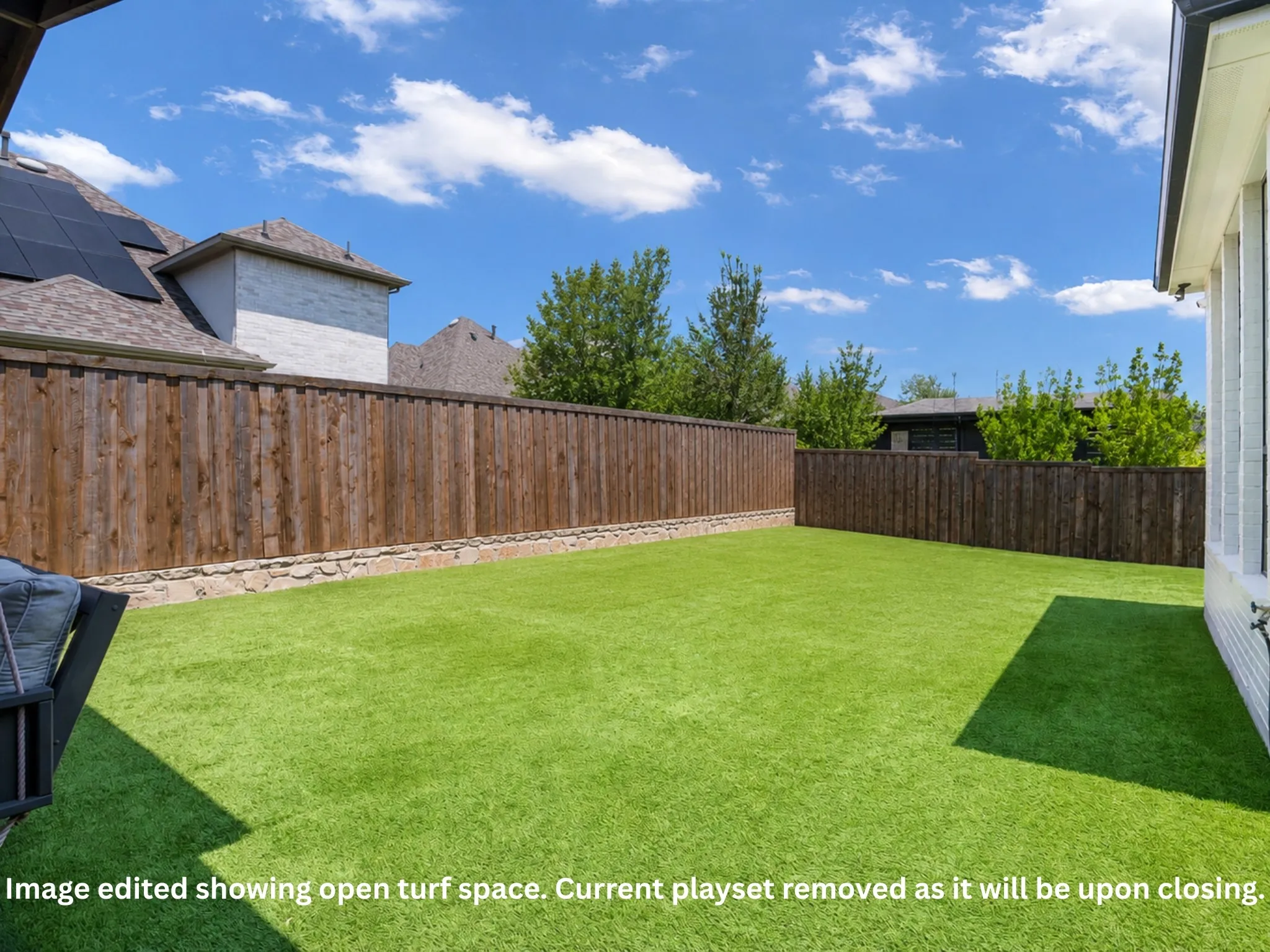 Expansive backyard featuring artificial turf, a wood-paneled fence with a stone base, and a portion of the main residence with solar panels visible on the roof