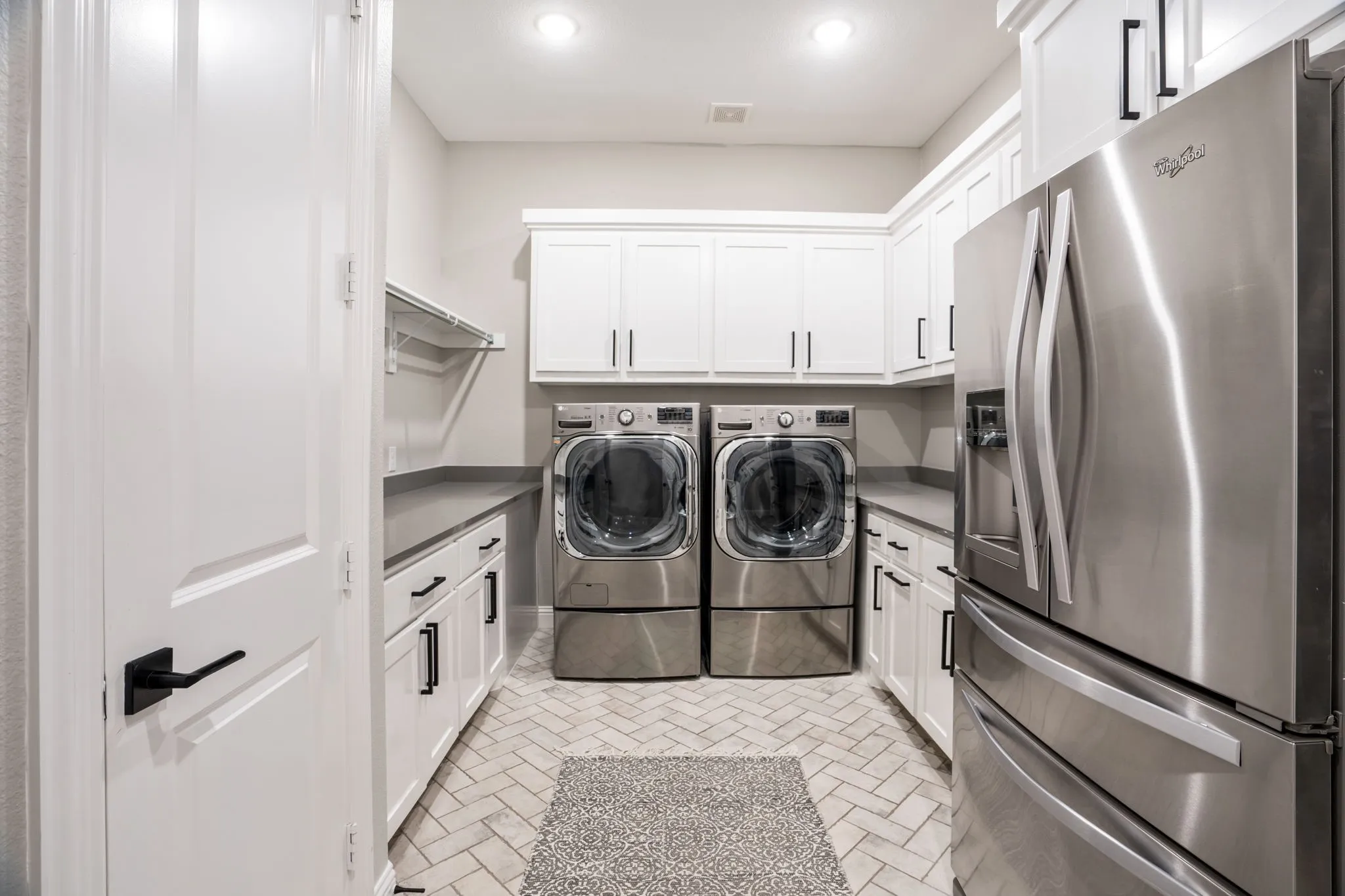 Dedicated laundry room featuring front-load washer and dryer with pedestal bases, white cabinetry with ample storage, stainless steel refrigerator, herringbone tile flooring, recessed lighting, and direct access to the home's main living areas
