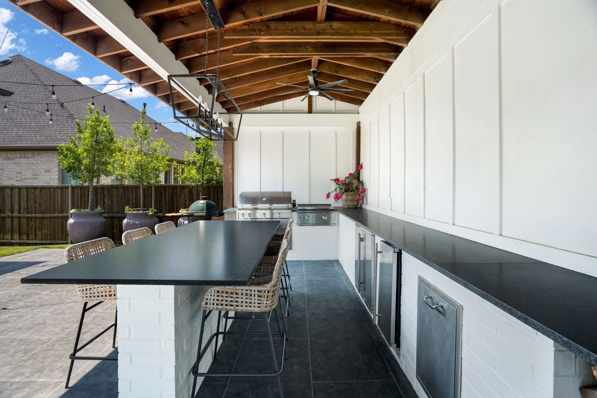 Sophisticated outdoor entertaining space featuring a custom black-finish dining table with seating for eight, professional-grade stainless steel grill and refrigeration, concrete flooring, and a covered pavilion with exposed wooden beam ceiling