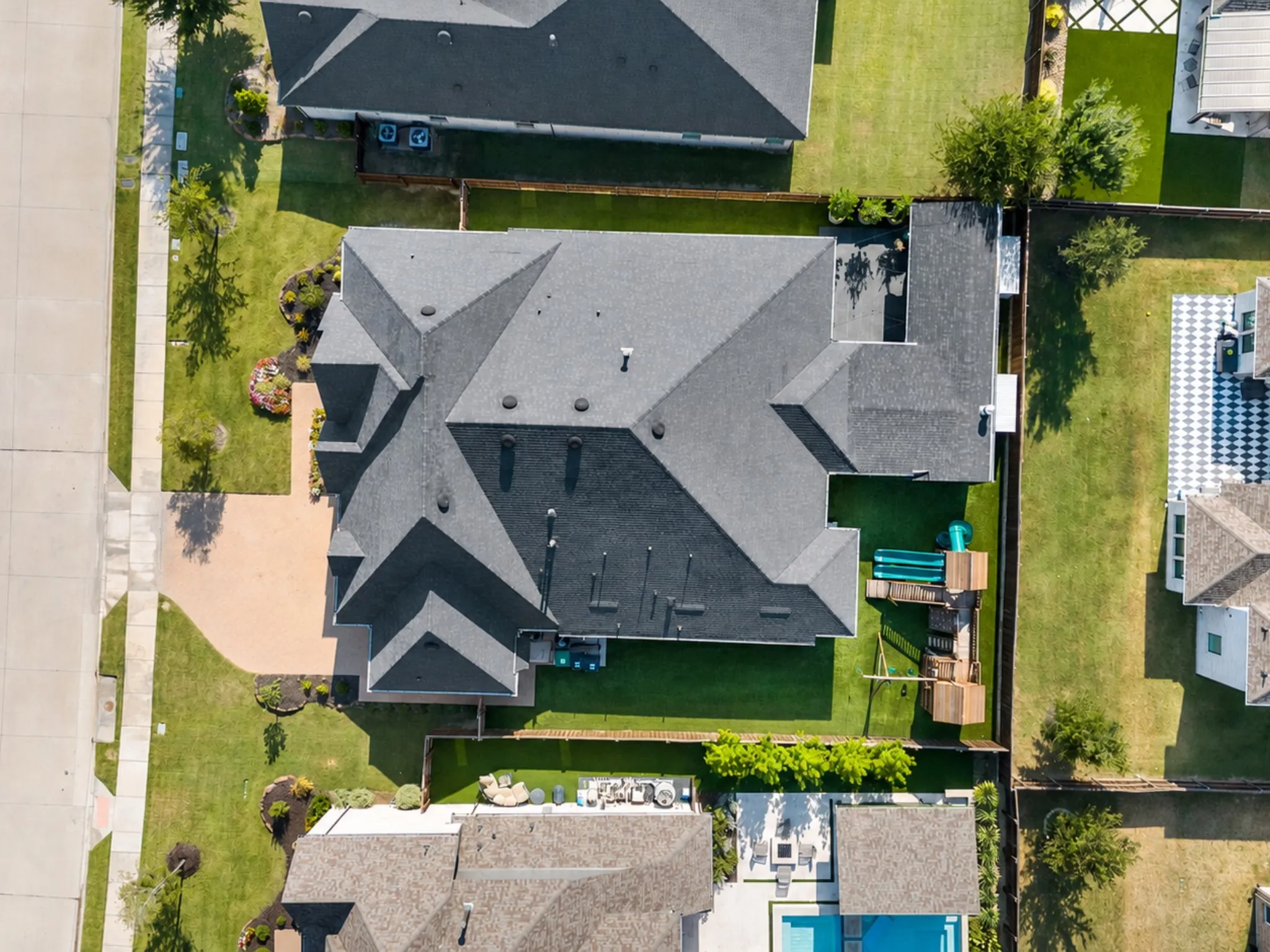 Aerial view showcasing a geometric multi-level roof design with dimensional gray asphalt shingles, complemented by manicured lawn, mature landscaping, and established trees throughout the property
