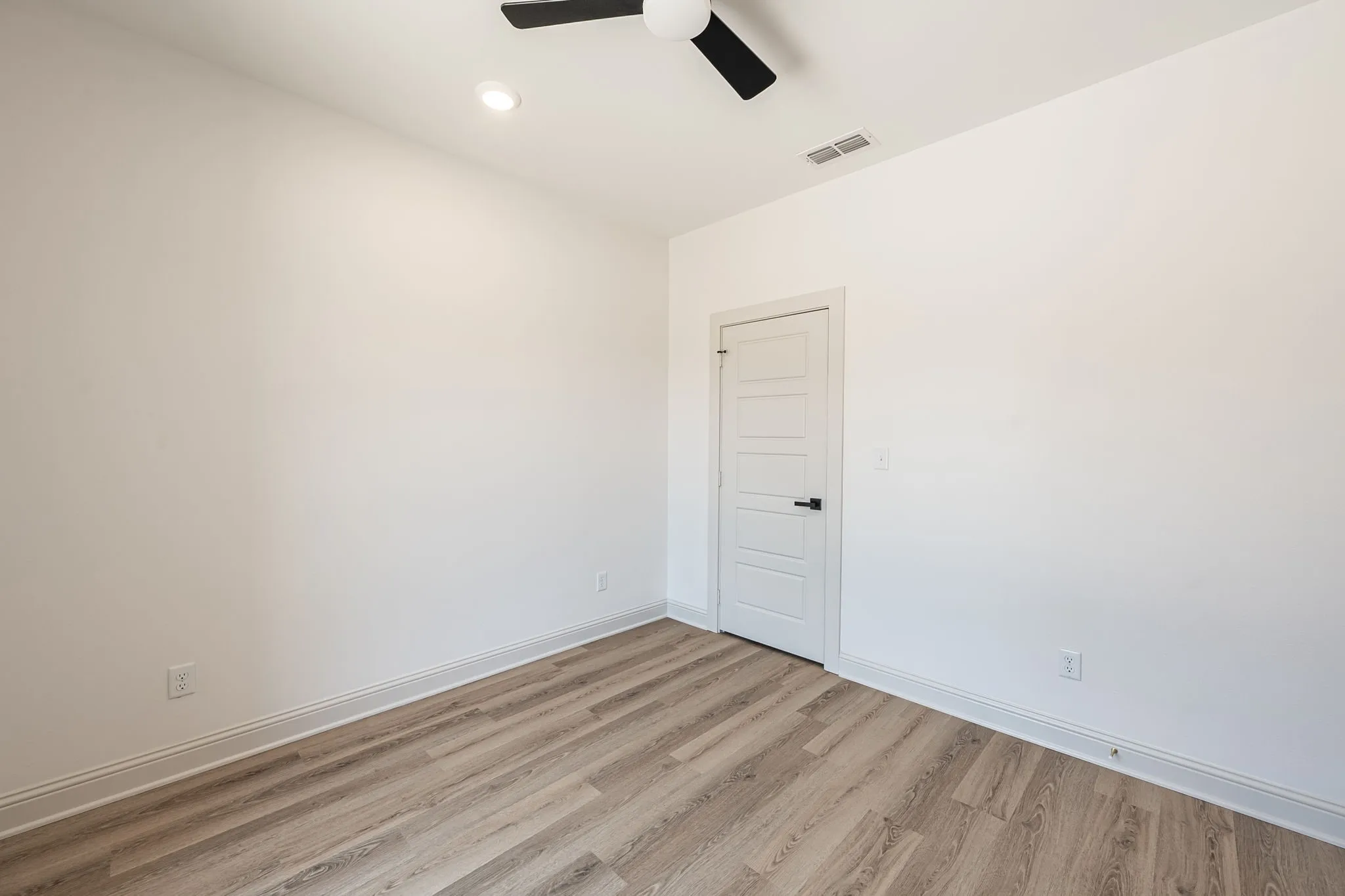 Recessed ceiling light, ceiling fan with black blades, white paneled door with black hardware, and wood-finish flooring