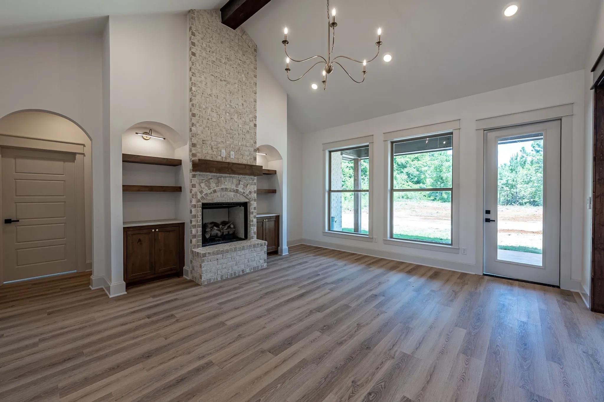 Bright open-concept living area featuring wood-finish flooring throughout, a striking stone fireplace with built-in shelving and lower cabinetry, arched architectural details, and abundant natural light from triple glass doors and multiple windows