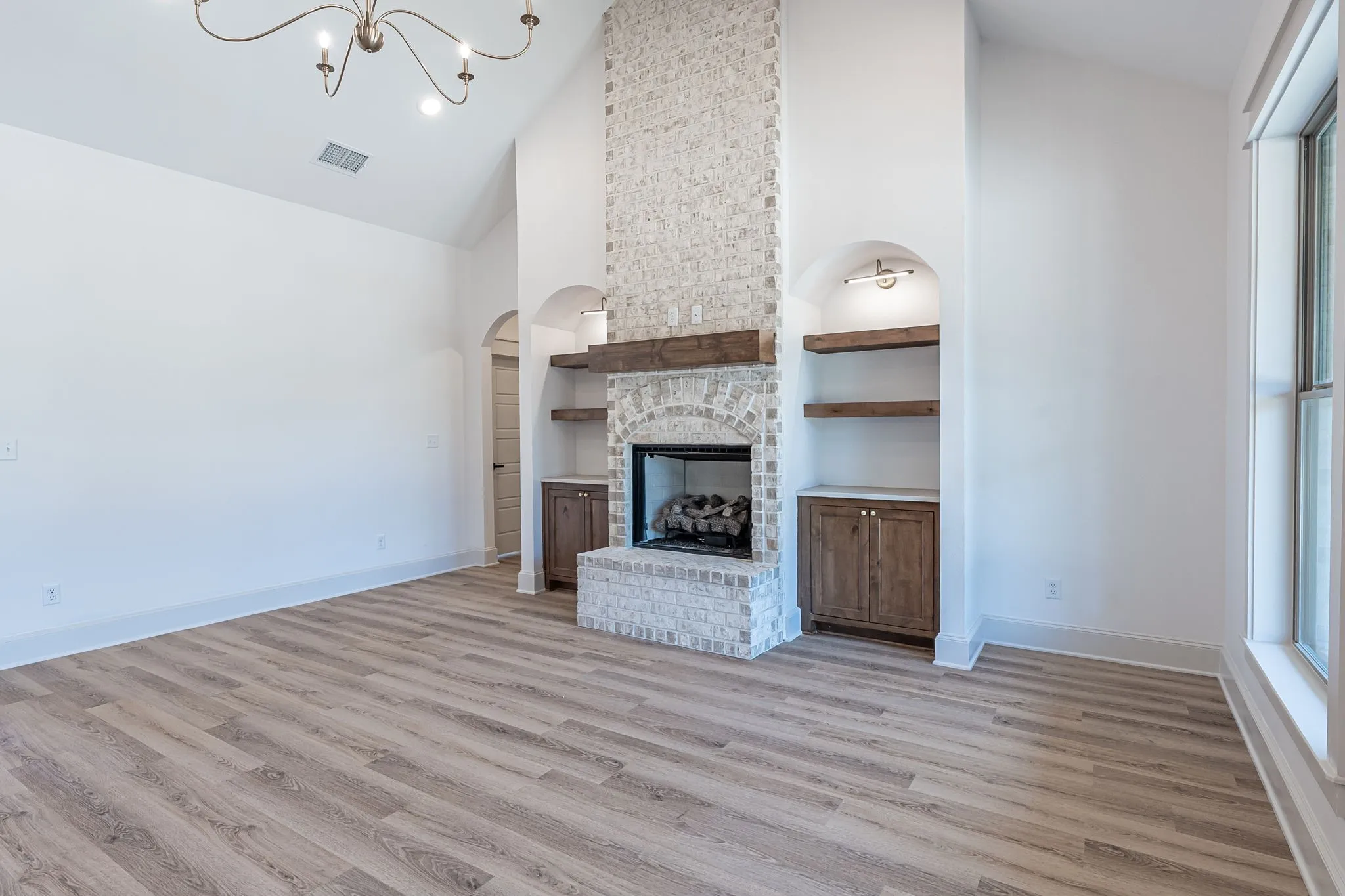 Vaulted ceiling with an ornate chandelier, fireplace with brick surround, built-in shelving and cabinetry, and wood-finish flooring