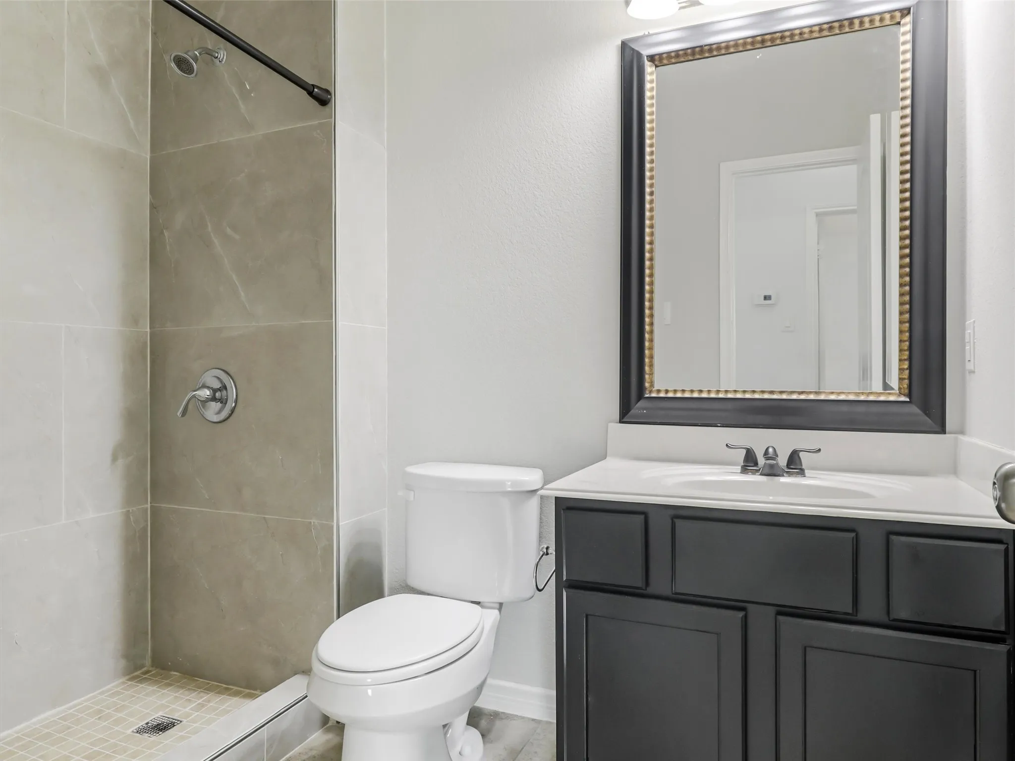 Full bathroom featuring a walk-in shower with large format tile surround, a white vanity with dark cabinetry, a framed mirror, and chrome fixtures
