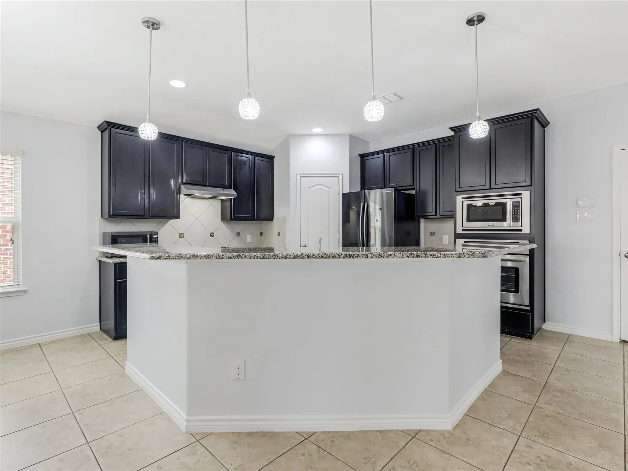 Kitchen featuring dark wood cabinetry, light-toned tile flooring, granite-style countertops, stainless steel appliances, and two decorative pendant lights