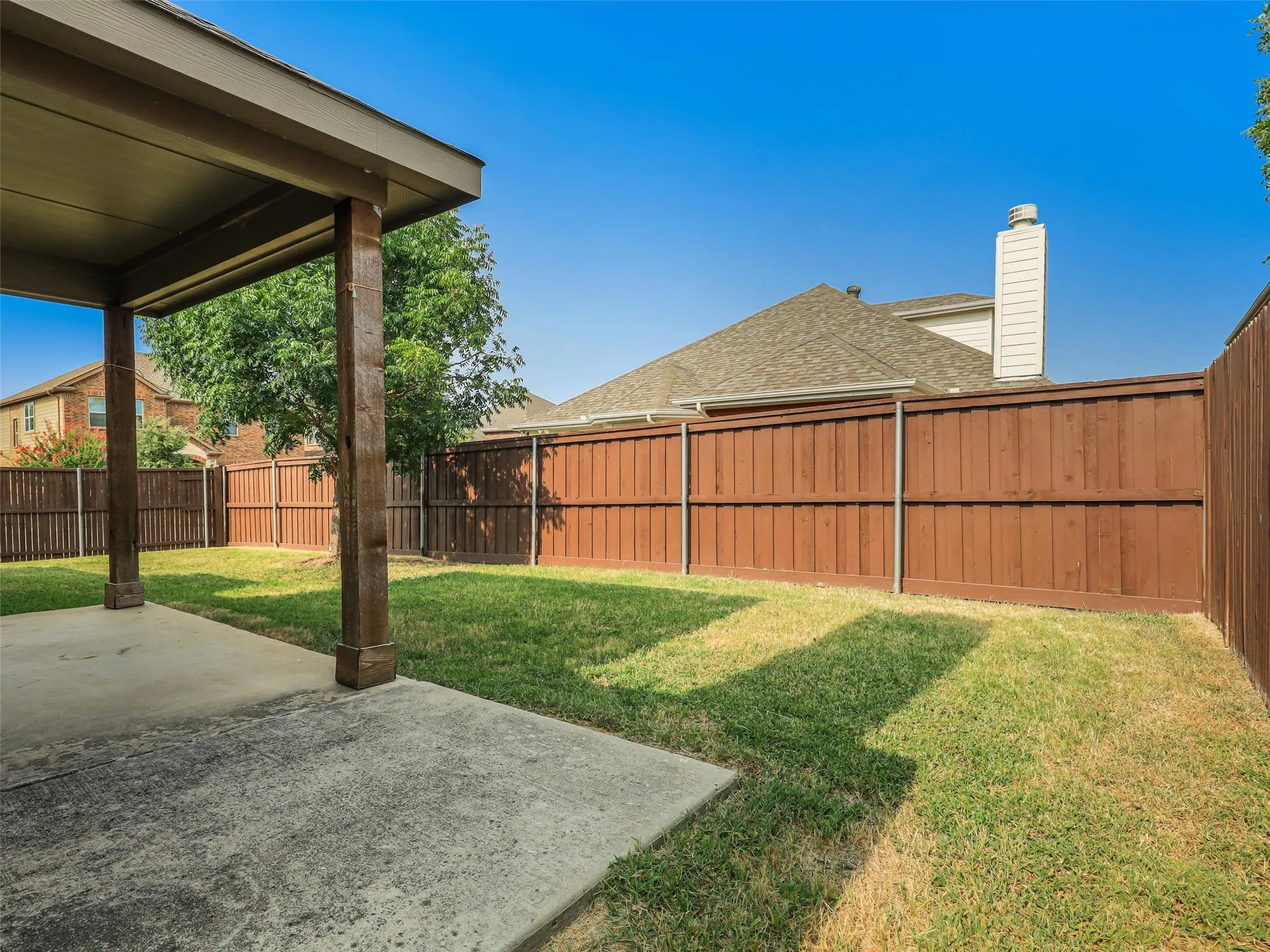 Expansive backyard featuring a covered patio, concrete paving, a manicured lawn, mature trees, and a privacy fence