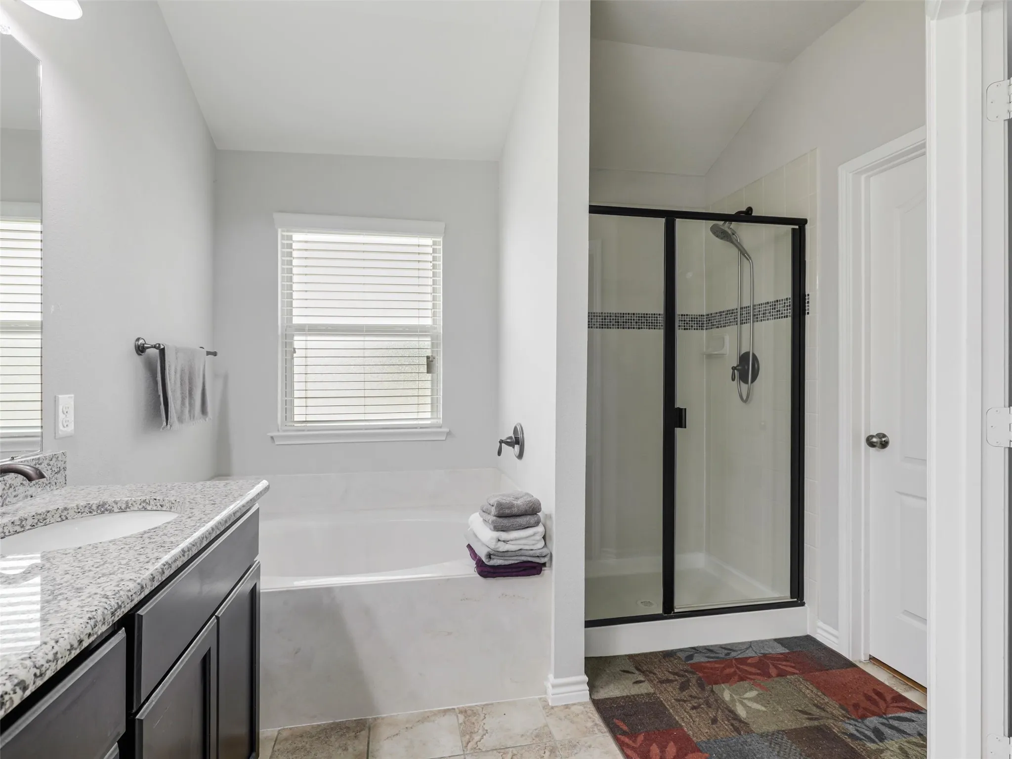 Bathroom featuring a built-in tub, separate shower with framed glass enclosure, vanity with granite-finish countertop, and a window with blinds