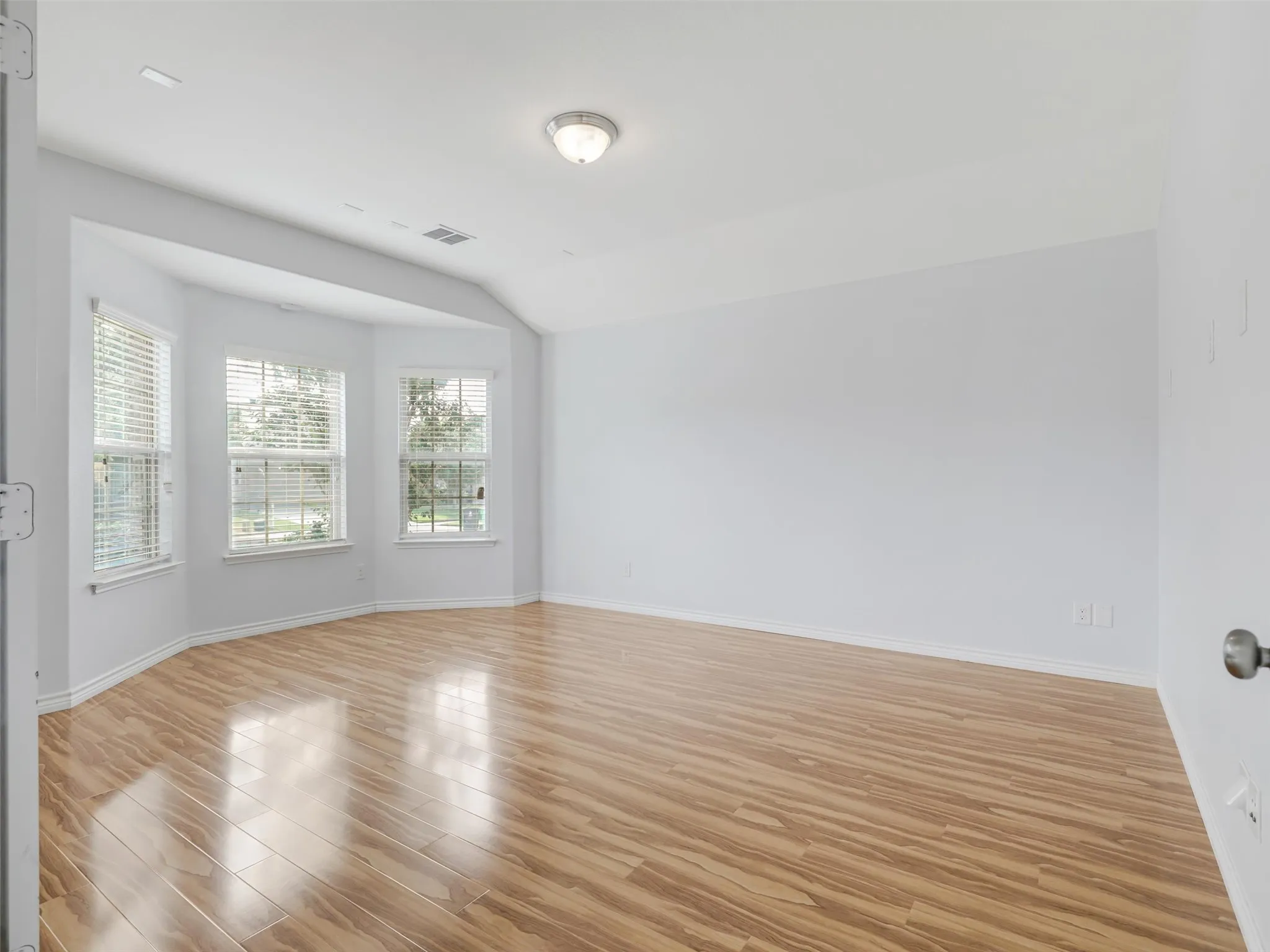 Spacious room with wood-finish flooring, featuring a bay window with three individual windows and blinds, neutral wall paint, and a semi-flush ceiling light fixture