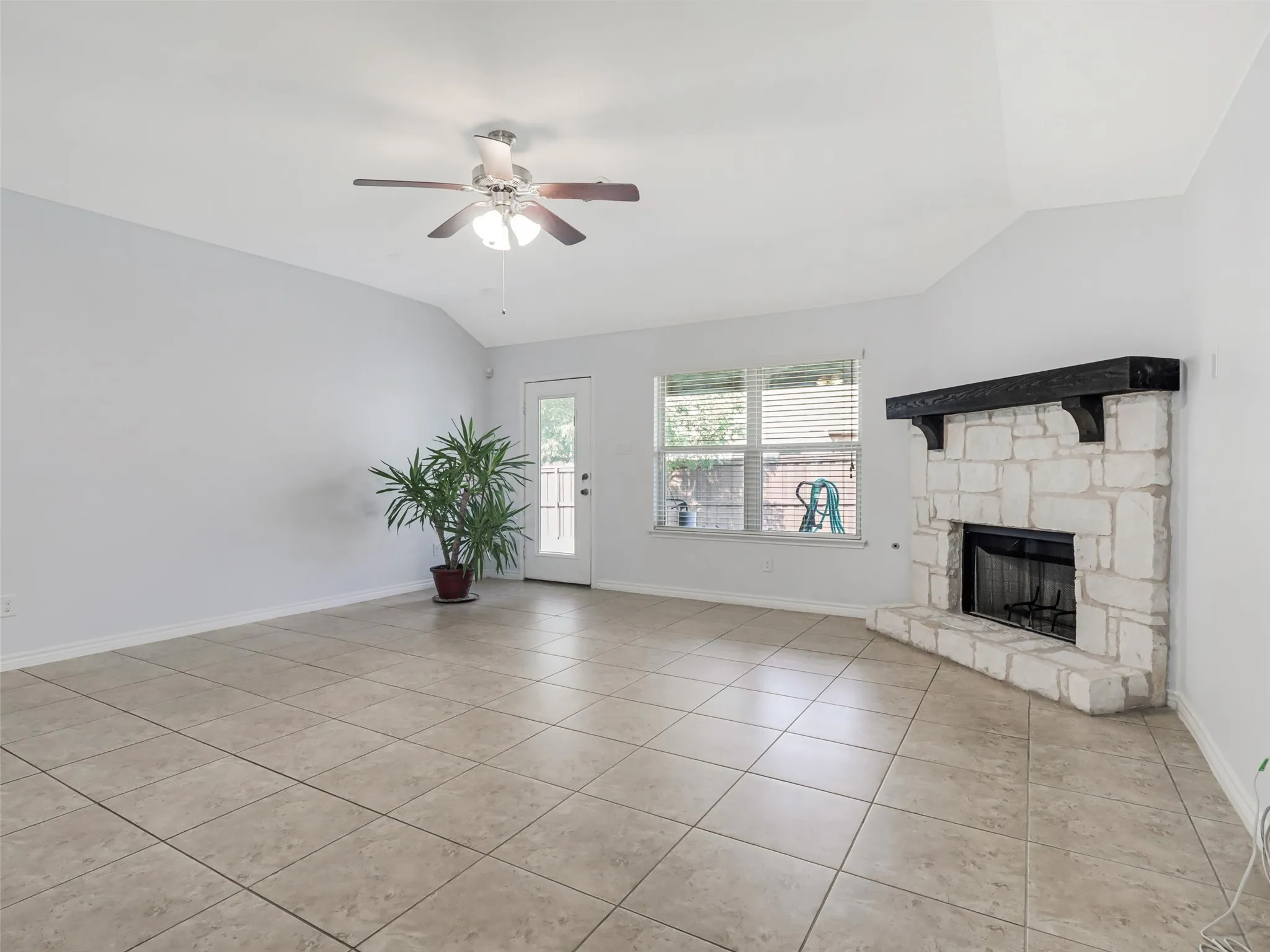 Spacious living area featuring a corner stone-clad fireplace with a dark wood mantel, vaulted ceiling, and ceramic tile flooring