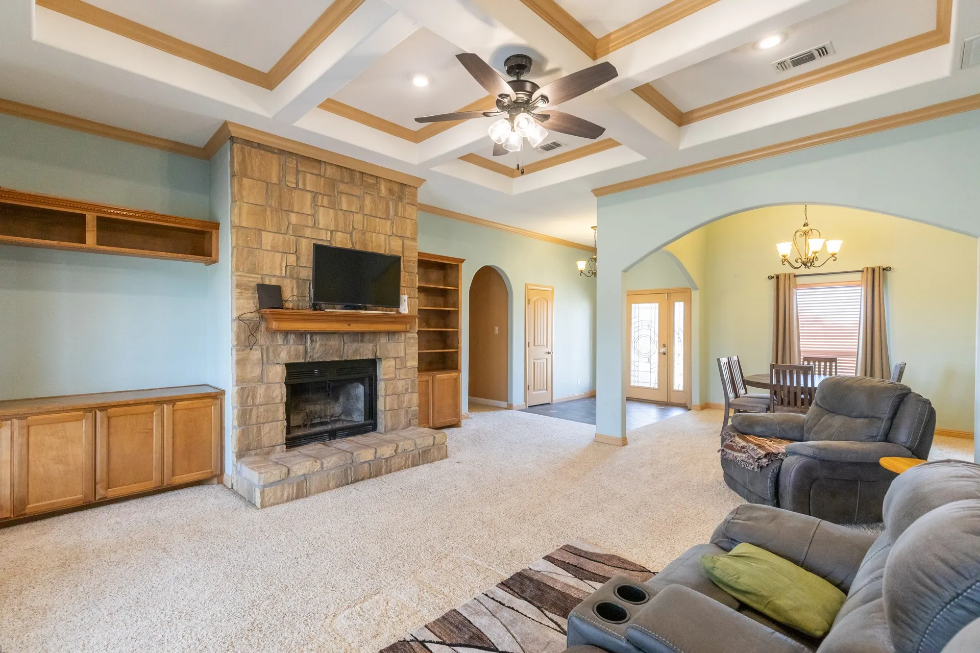 Spacious great room featuring a wood-burning fireplace with stone surround and custom wood cabinetry, coffered ceiling with ceiling fan, and recessed lighting throughout