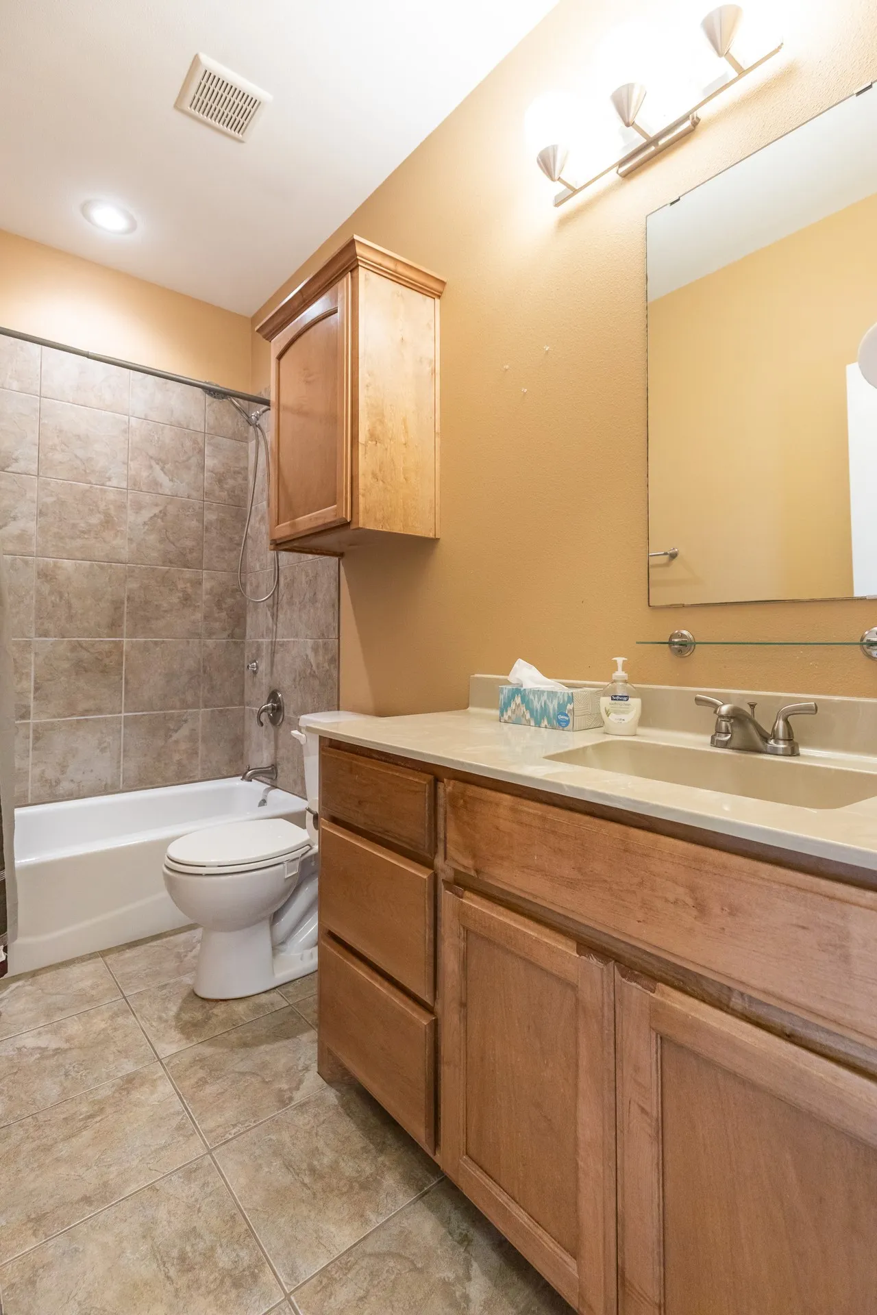 Spa-inspired master bathroom featuring warm wood cabinetry with ample storage, a solid-surface countertop with dual-sink vanity, large framed mirror with integrated lighting, and an enclosed shower-tub combination with tile surround