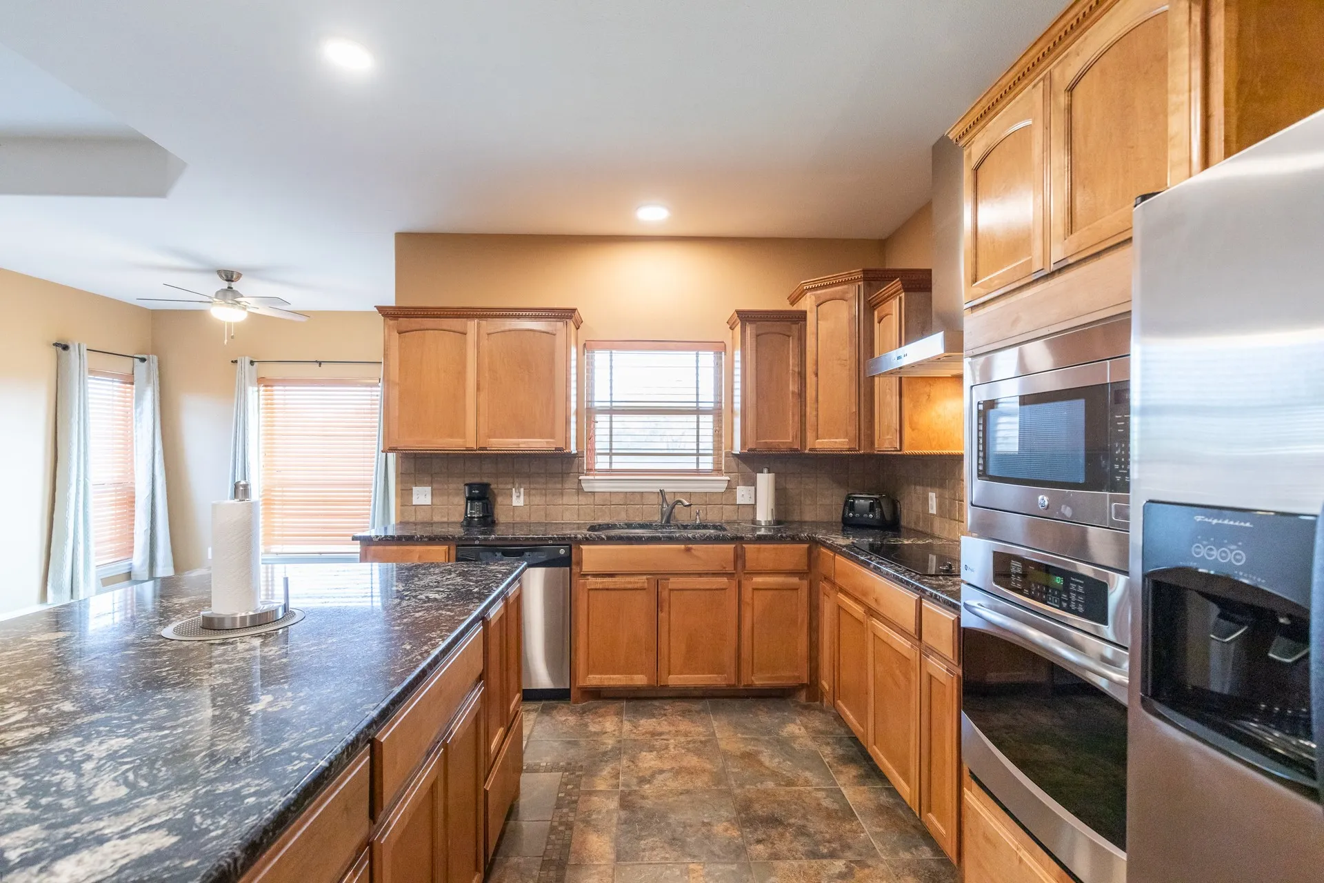Kitchen featuring a substantial island with a dark stone countertop, wood cabinetry, stainless steel appliances, tile flooring, and a tile backsplash