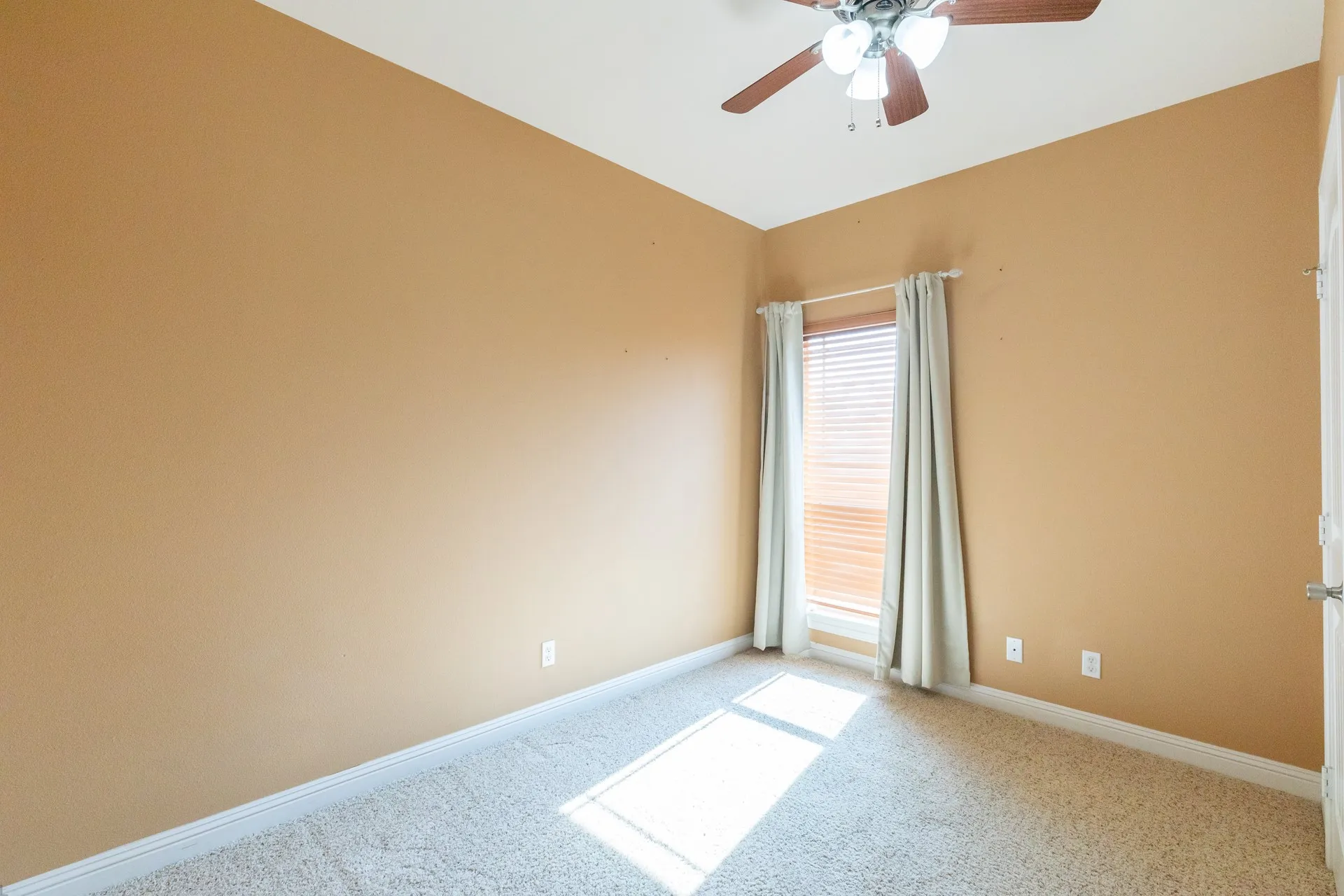 Bright and spacious bedroom featuring warm golden-toned walls, neutral carpet flooring, and abundant natural light from a full-length window with plantation shutters