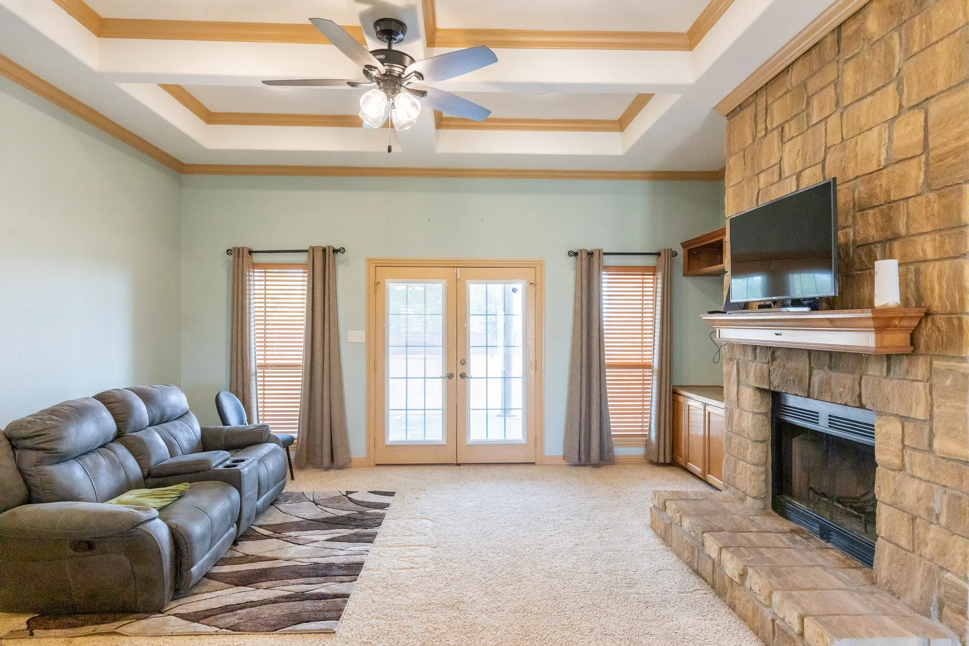 Spacious living room featuring a coffered ceiling with wood trim, ceiling fan with integrated lighting, and a brick fireplace with built-in shelving and mounted television