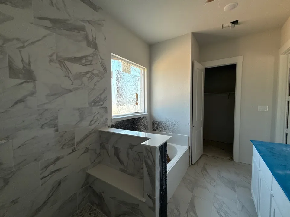 Spa-inspired primary bathroom featuring elegant marble-look tile throughout walls and flooring, a soaking tub with integrated surround, a wide vanity with ample counter space, and a large window flooding the space with natural light
