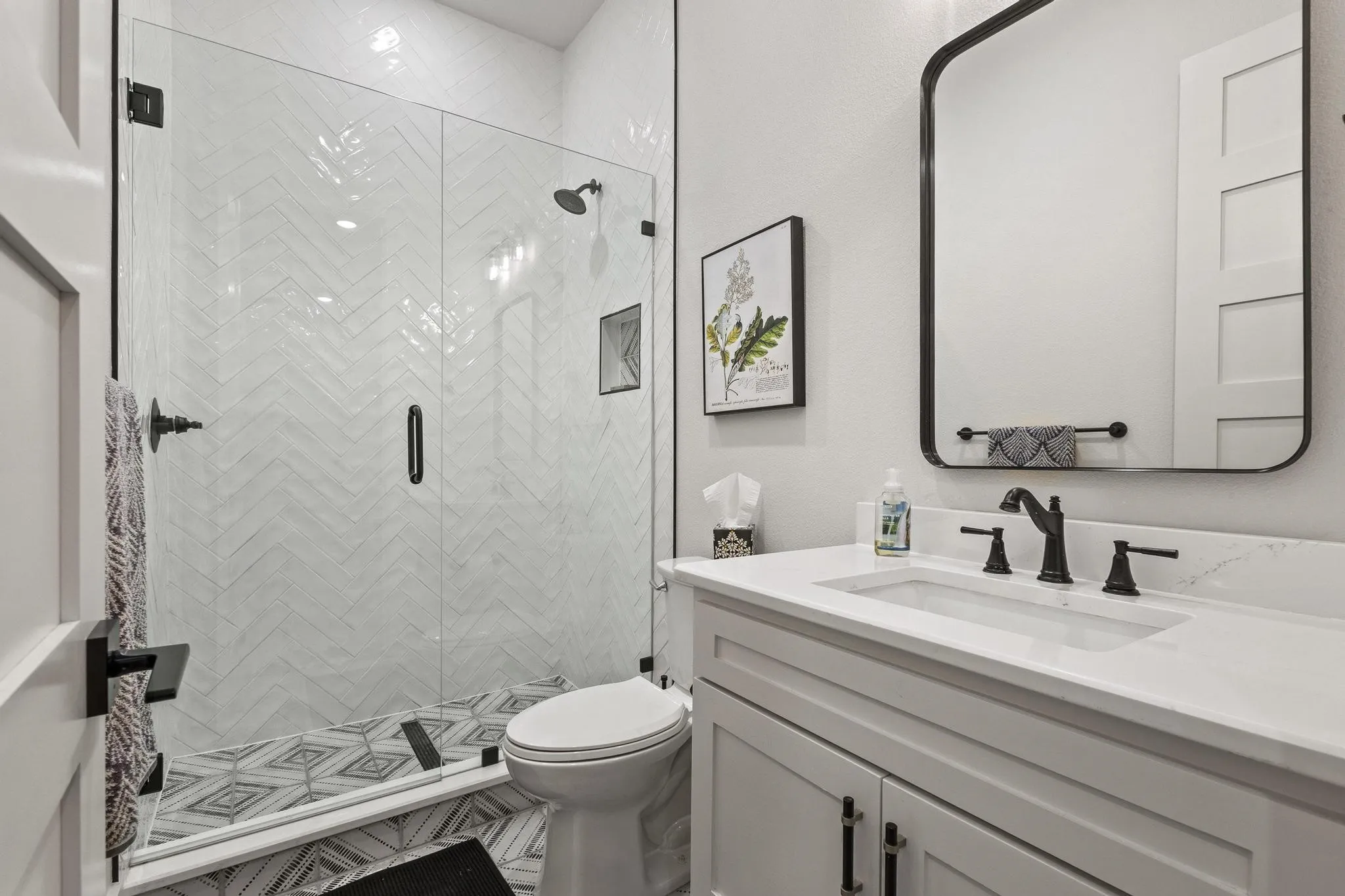 Modern bathroom featuring a glass-enclosed shower with white herringbone tile, a patterned tile floor, a white vanity with a rectangular sink, a white countertop, and matte black fixtures