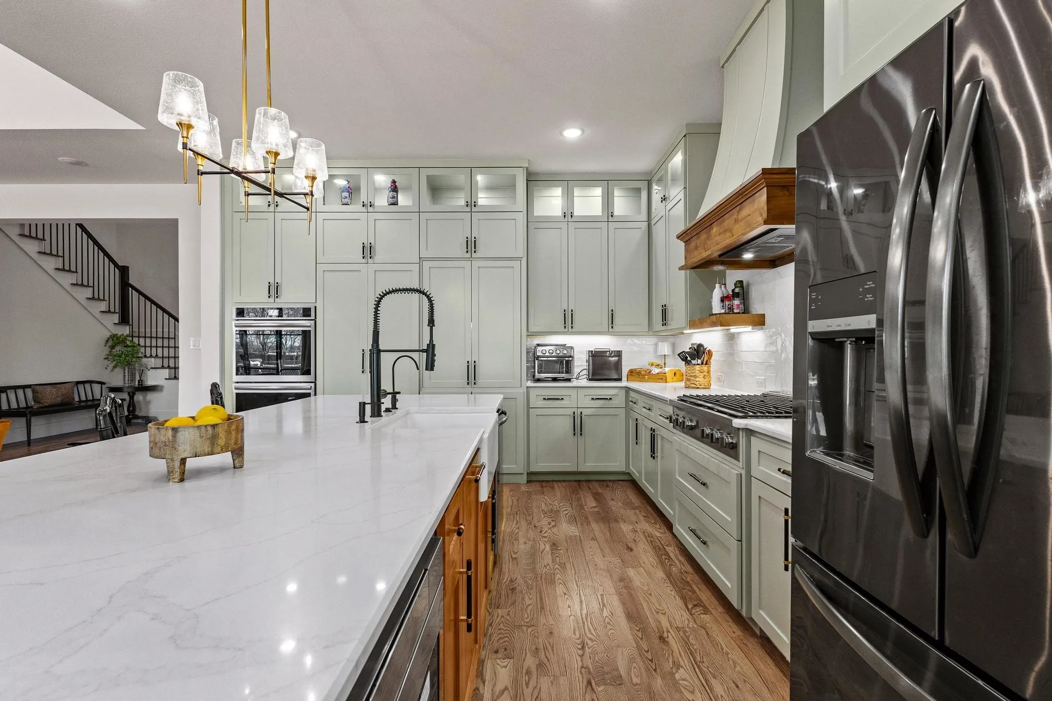 Gourmet kitchen featuring a substantial island with a white solid surface countertop, light green cabinetry, hardwood flooring, and stainless steel appliances