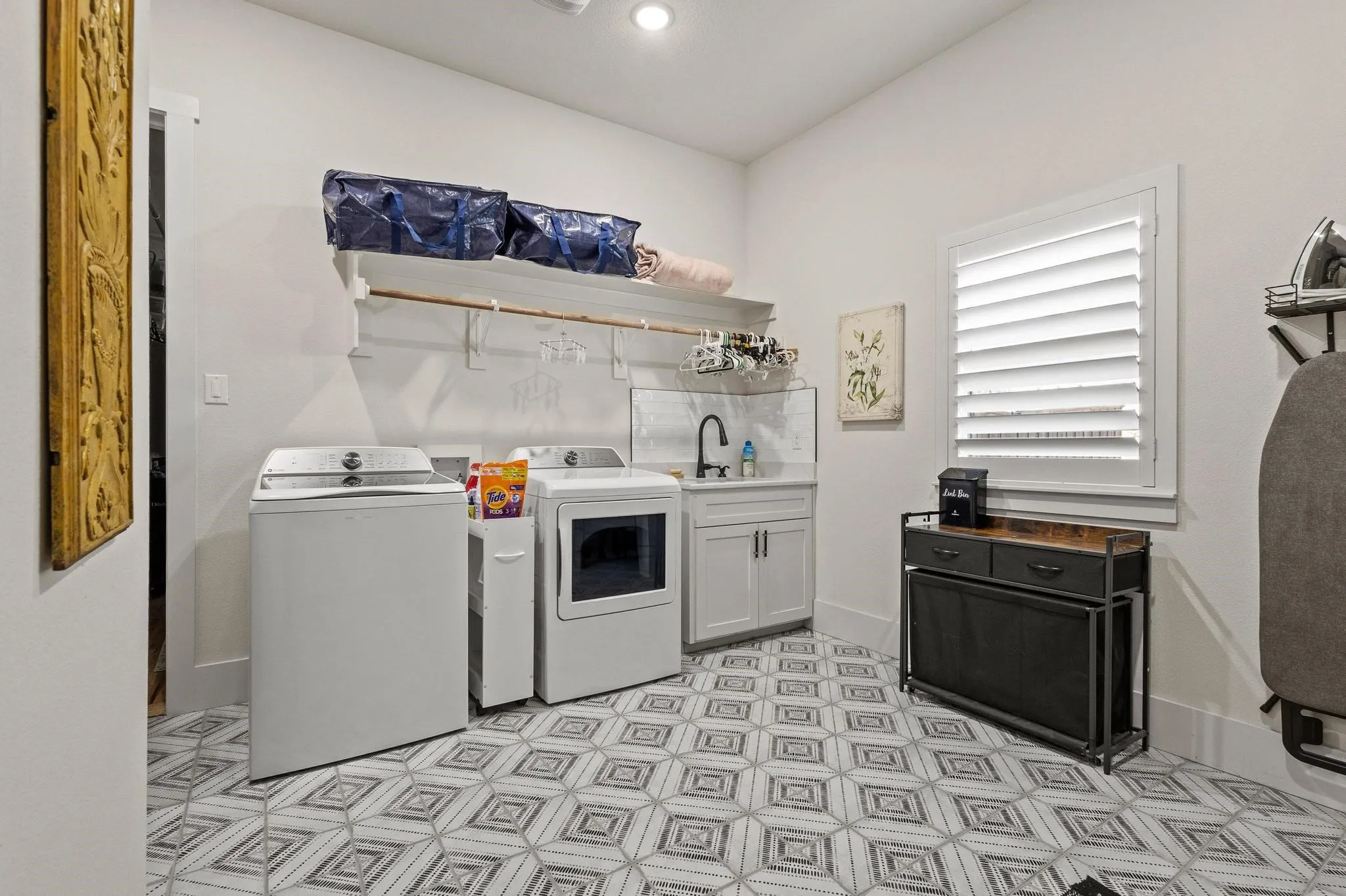 Laundry room featuring patterned tile flooring, a utility sink with a matte black faucet, and white plantation shutters on the window