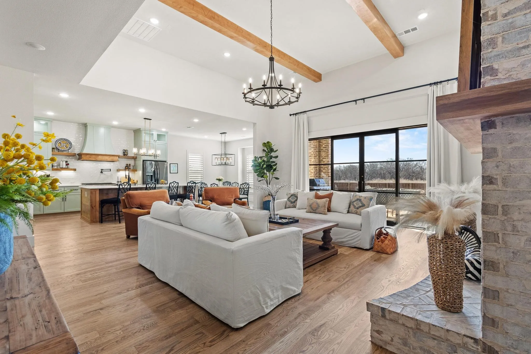 Spacious open-concept living area featuring exposed wood beams, wood-finish flooring, a stone fireplace, and expansive rear windows