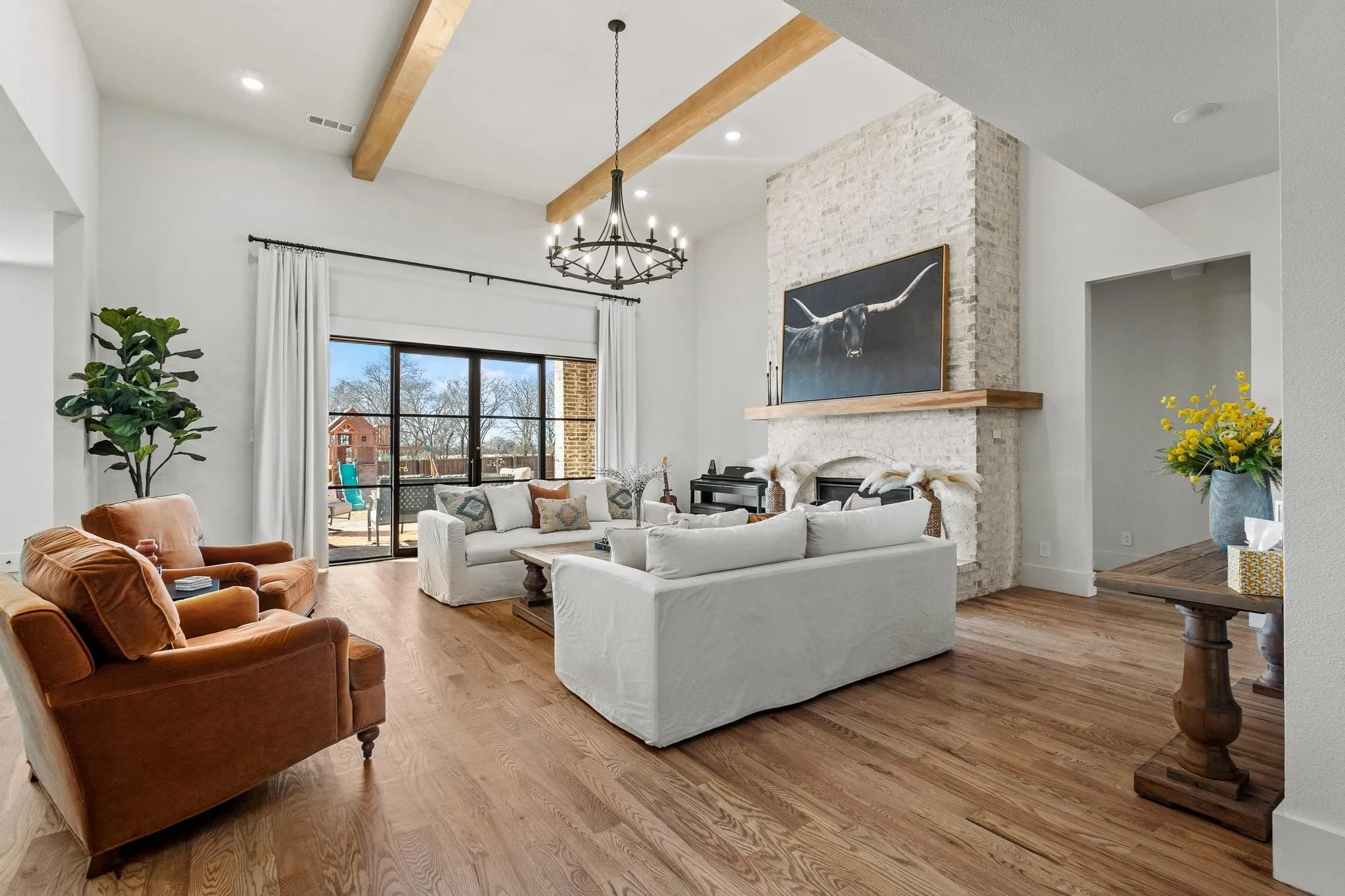Spacious living area featuring high vaulted ceilings with exposed wood beams, a prominent brick fireplace with a wood mantel, wide plank wood-finish flooring, and large sliding glass doors providing access to exterior