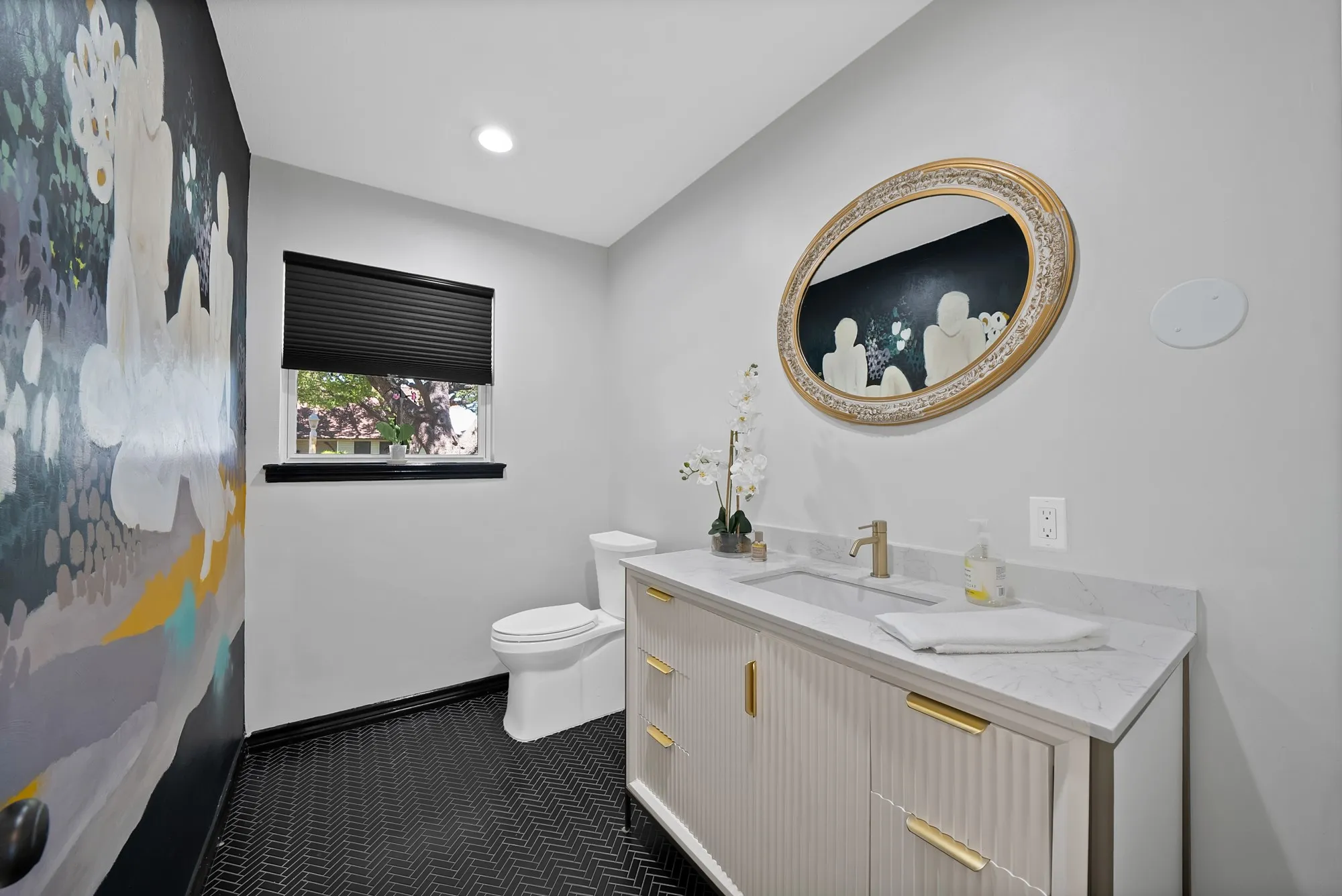 Herringbone tile flooring, vanity with gold-tone hardware, built-in sink, gold-tone faucet, and oval mirror with textured frame