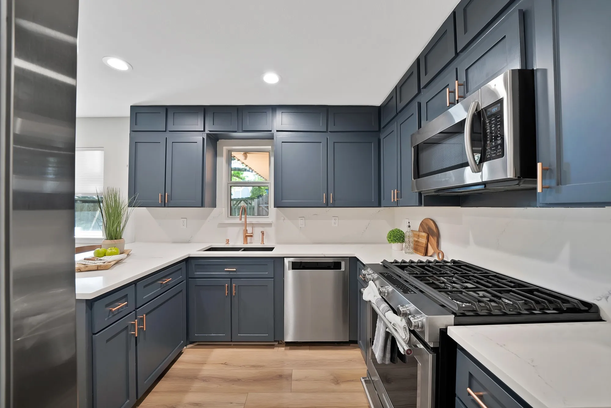 White countertops, recessed lighting, stainless steel appliances, wood-finish flooring, and blue shaker-style cabinetry with rose gold hardware