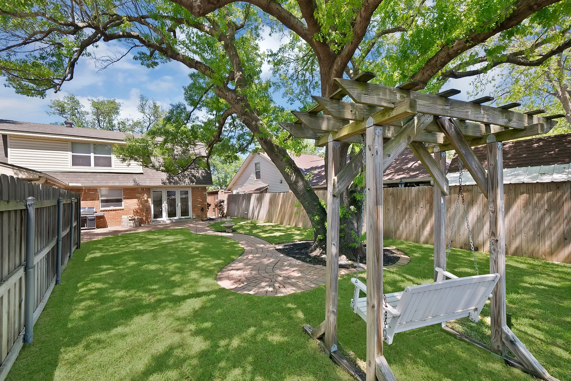 Spacious backyard featuring a manicured lawn, paved walkway, mature tree, and pergola with swing